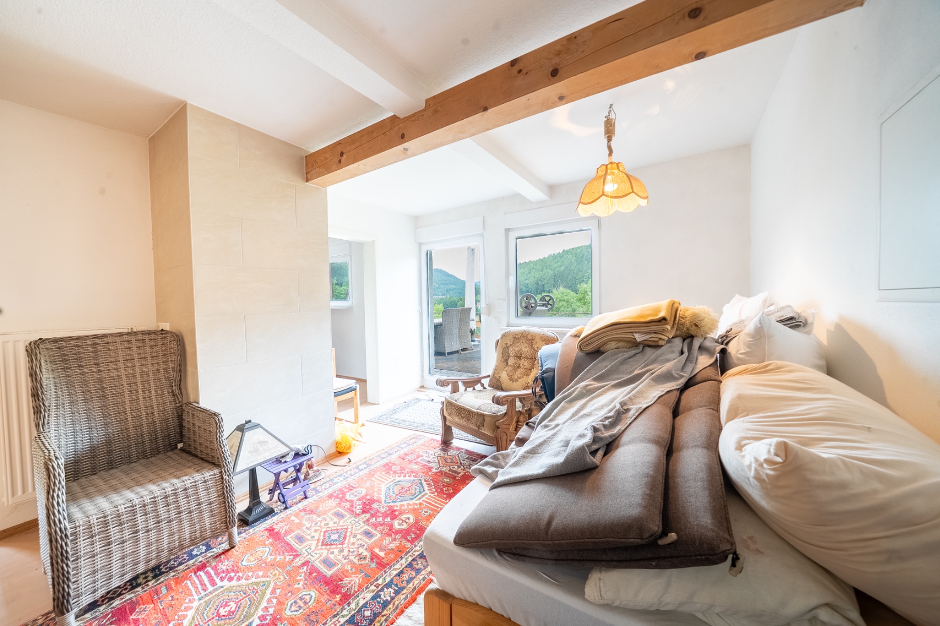 Helles Zimmer mit Bett, Sessel, Teppich und großem Fenster, das auf grüne Berge blickt.