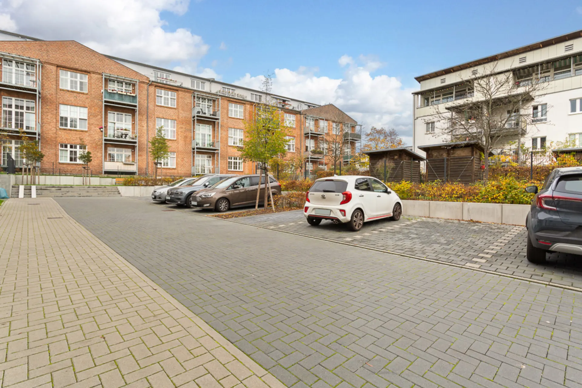 Mehrere Autos parken auf gepflastertem Parkplatz vor Wohngebäuden, einige Bäume im Hintergrund.