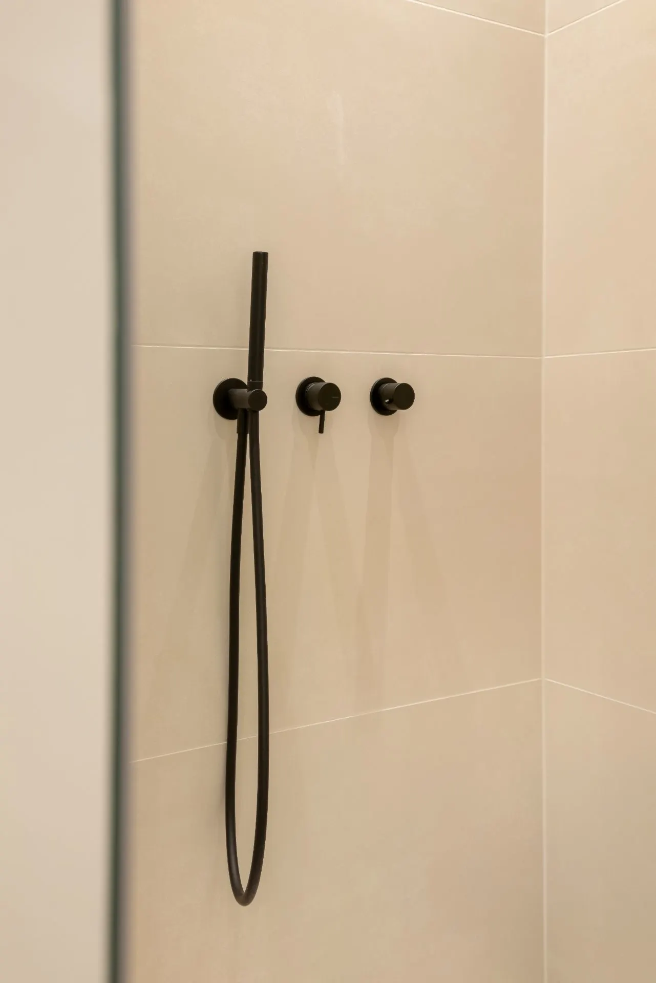 Moderne Dusche mit schwarzer Handbrause und drei Bedienelementen an beiger Wand.