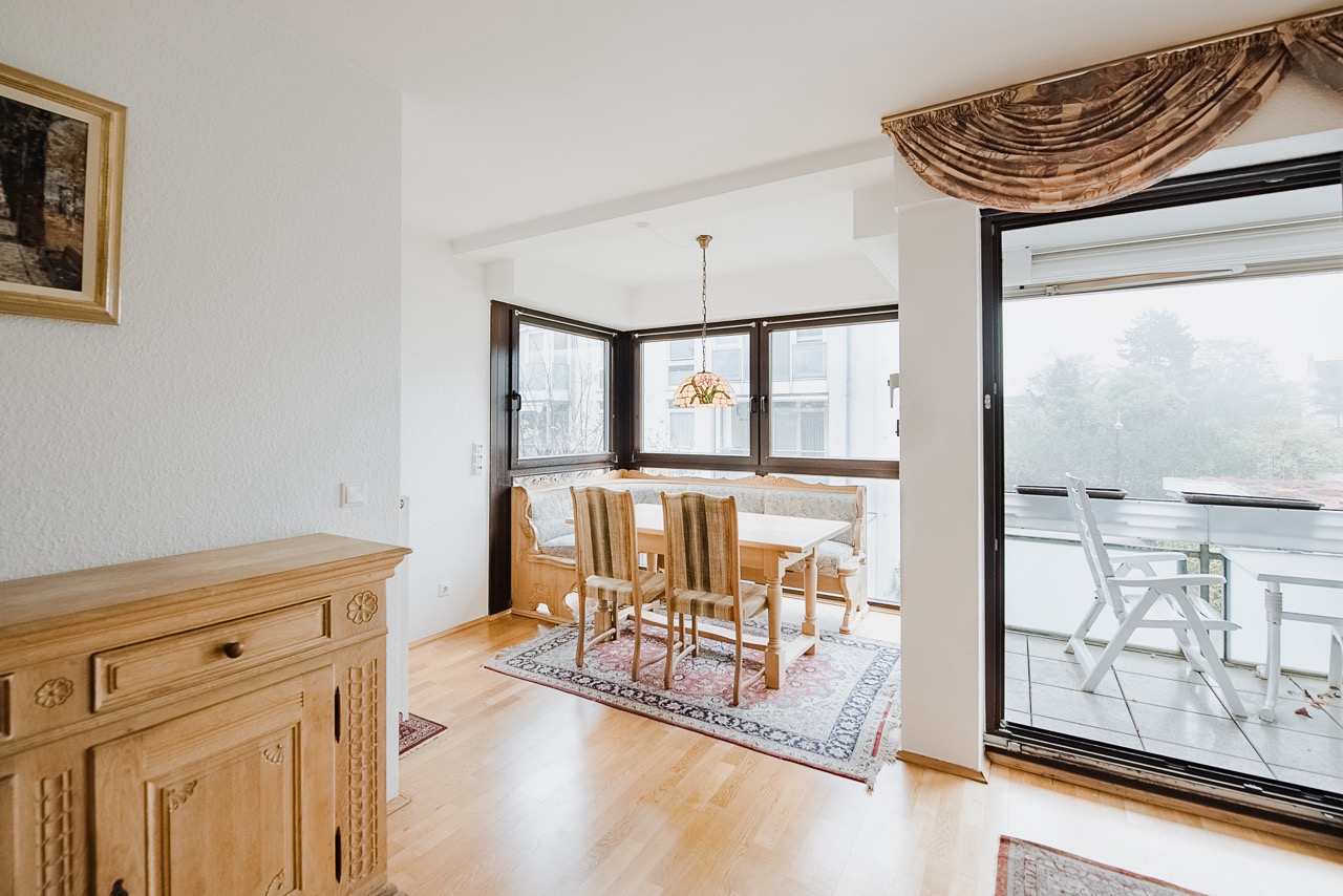 Essbereich mit Holztisch, Stühlen und Eckbank neben großen Fenstern, Zugang zu Balkon mit Stühlen.