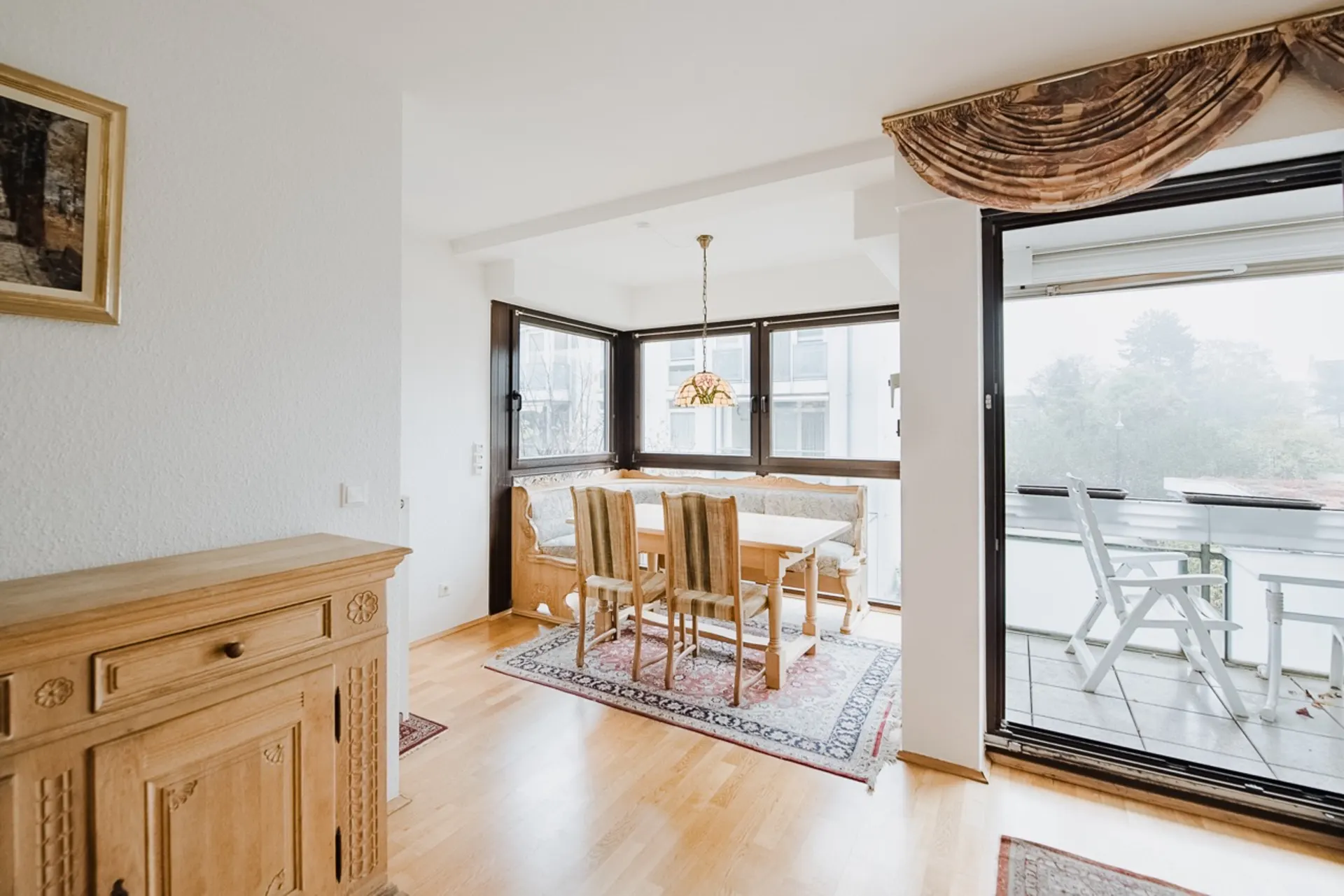 Essbereich mit Holztisch, Stühlen und Eckbank neben großen Fenstern, Zugang zu Balkon mit Stühlen.