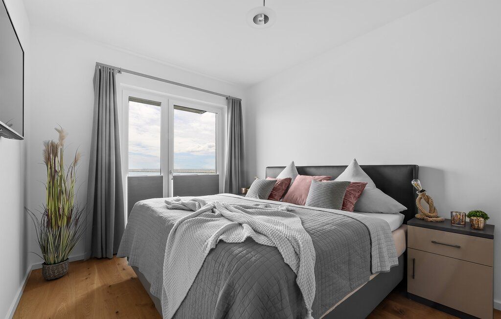 Schlafzimmer mit Doppelbett, grauen Vorhängen, Nachttisch und Dekoration, Tageslicht durch Fenster.