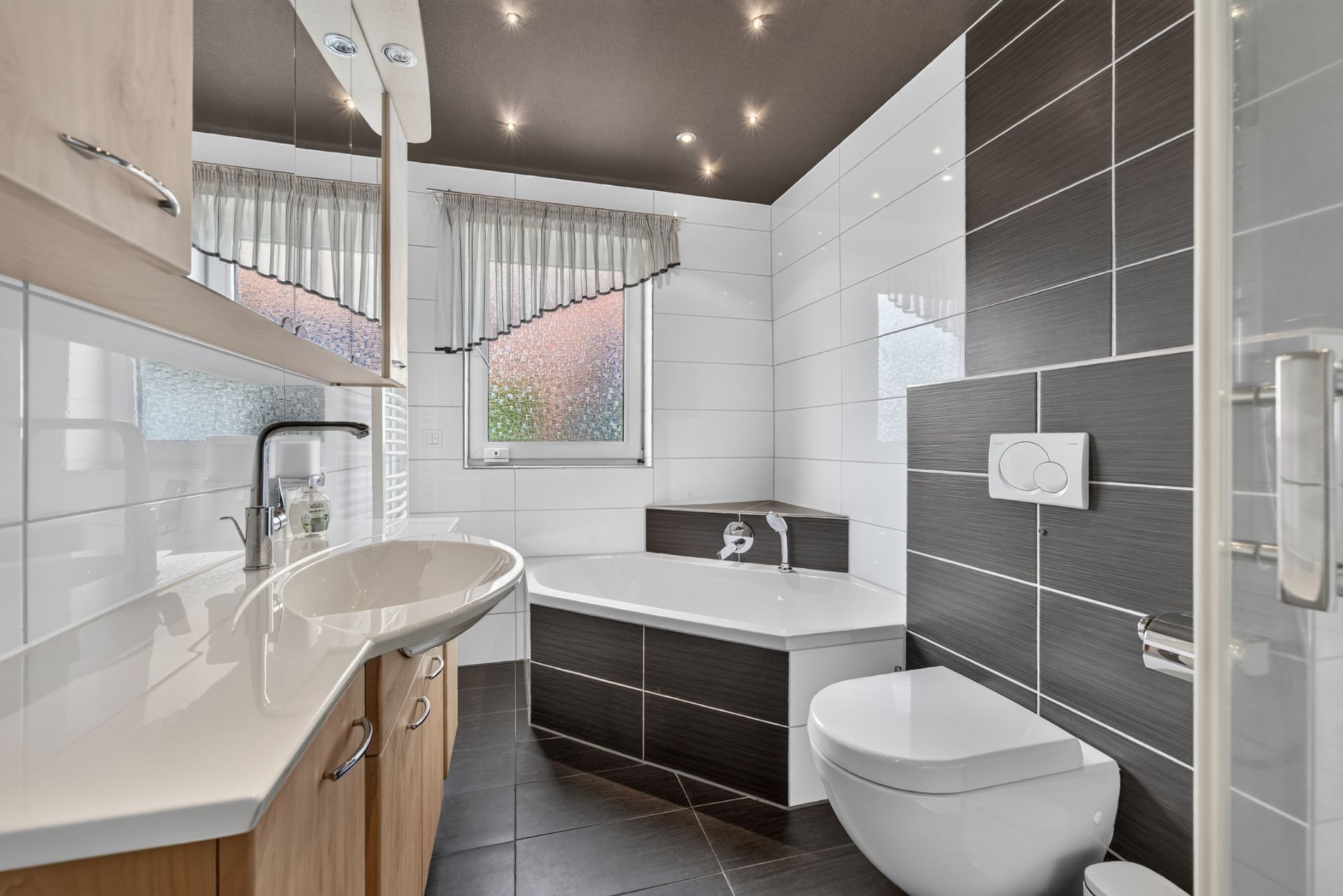 Modernes Badezimmer mit Badewanne, Waschbecken, Toilette und Fenster mit Vorhängen.