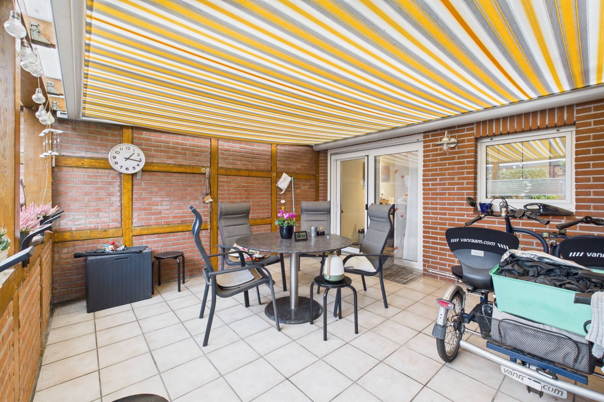 Überdachte Terrasse mit Tisch, Stühlen, Uhr an der Wand und Fahrrädern auf gefliestem Boden.