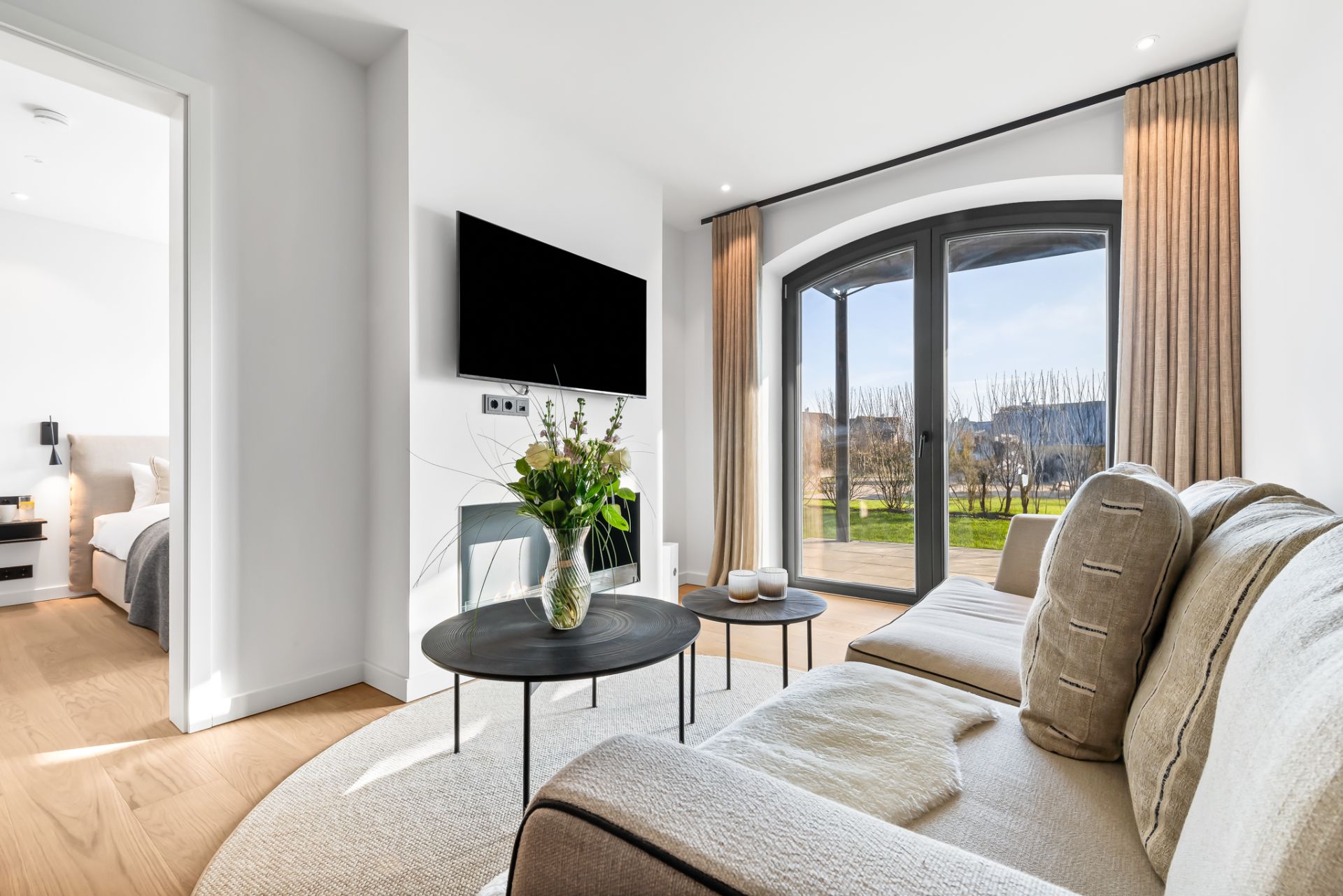 Helles Wohnzimmer mit Sofa, Couchtisch, Fernseher und großen Fenstern mit Blick ins Grüne.