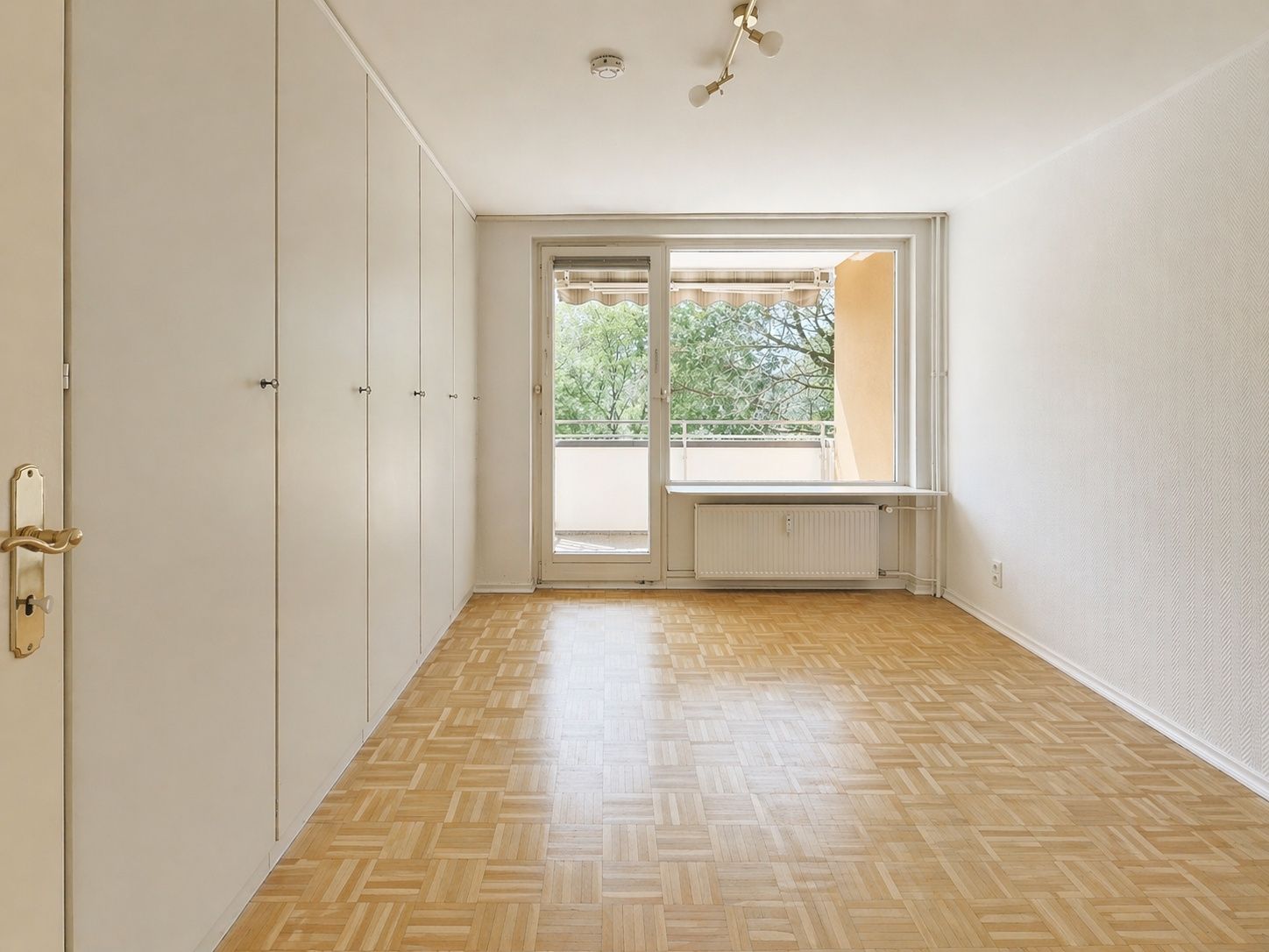 Heller Raum mit Parkettboden, Einbauschränken und Zugang zu einem Balkon mit Markise.