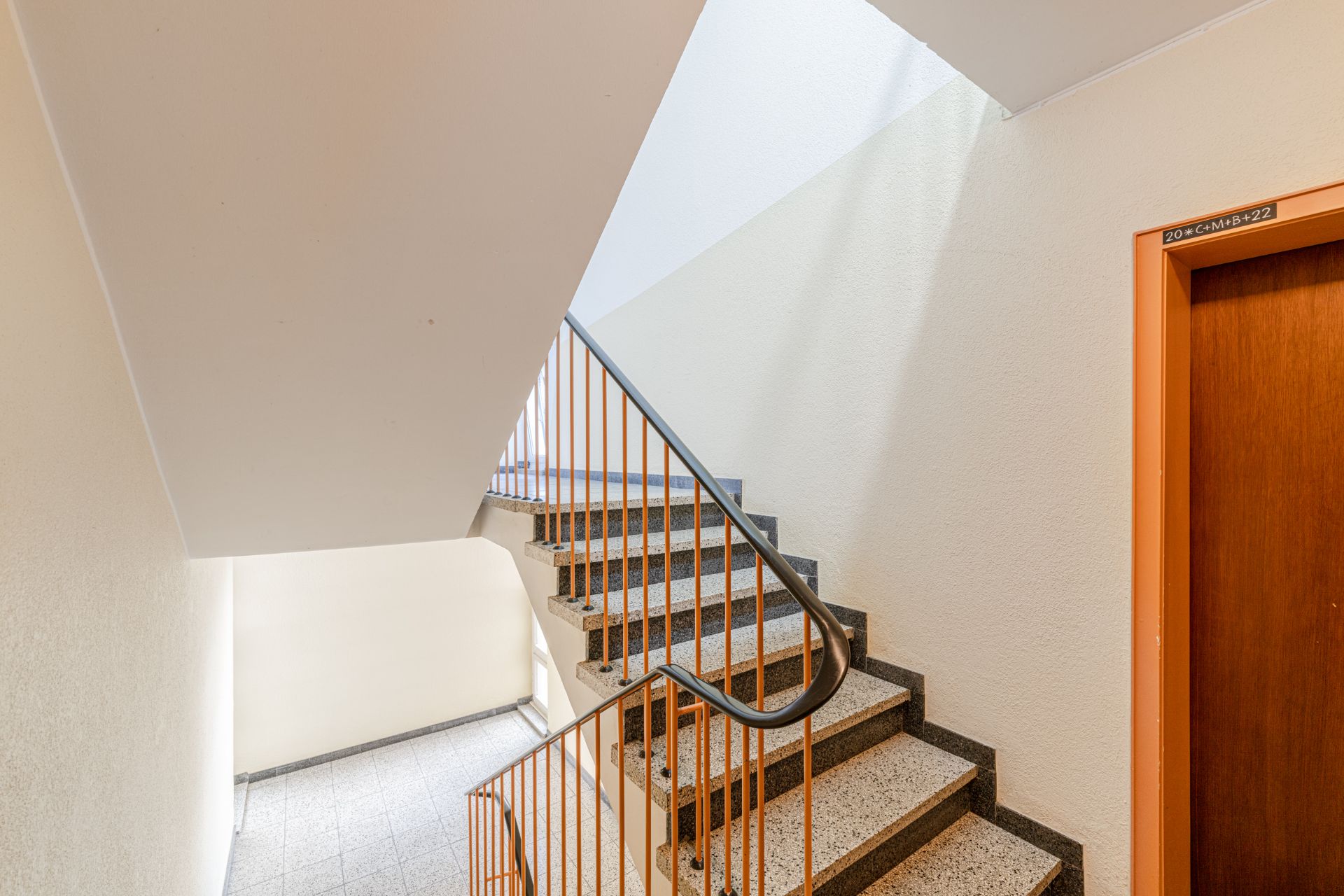 Helles Treppenhaus mit Marmorstufen, orangem Geländer und einer Holztür rechts.