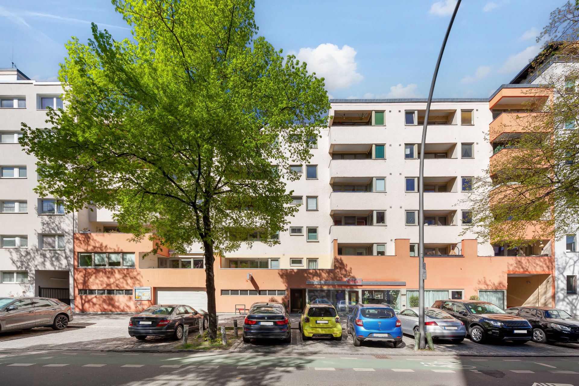 Mehrstöckiges Wohnhaus mit Balkonen, davor parken Autos und ein großer Baum steht auf dem Gehweg.