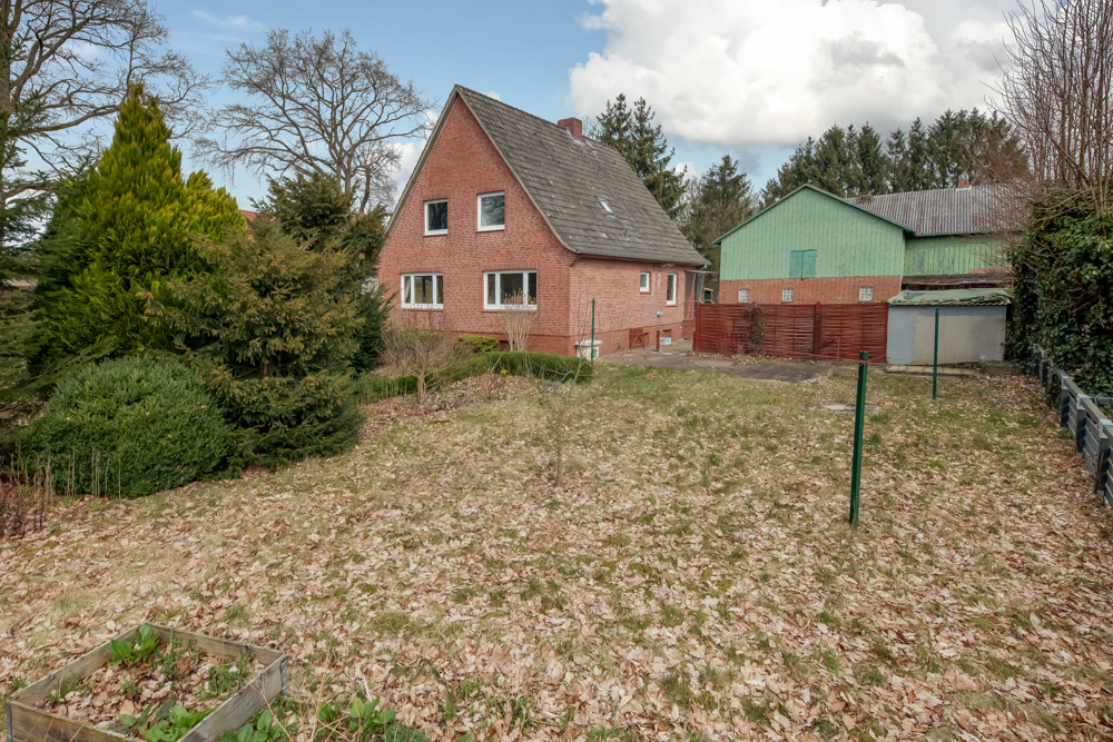 Backsteinwohnhaus mit Garten und Bäumen im Vordergrund, Nebengebäude im Hintergrund.