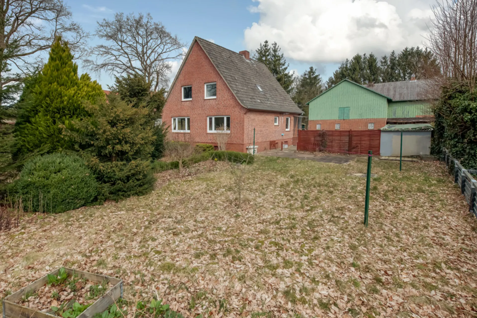 Backsteinwohnhaus mit Garten und Bäumen im Vordergrund, Nebengebäude im Hintergrund.
