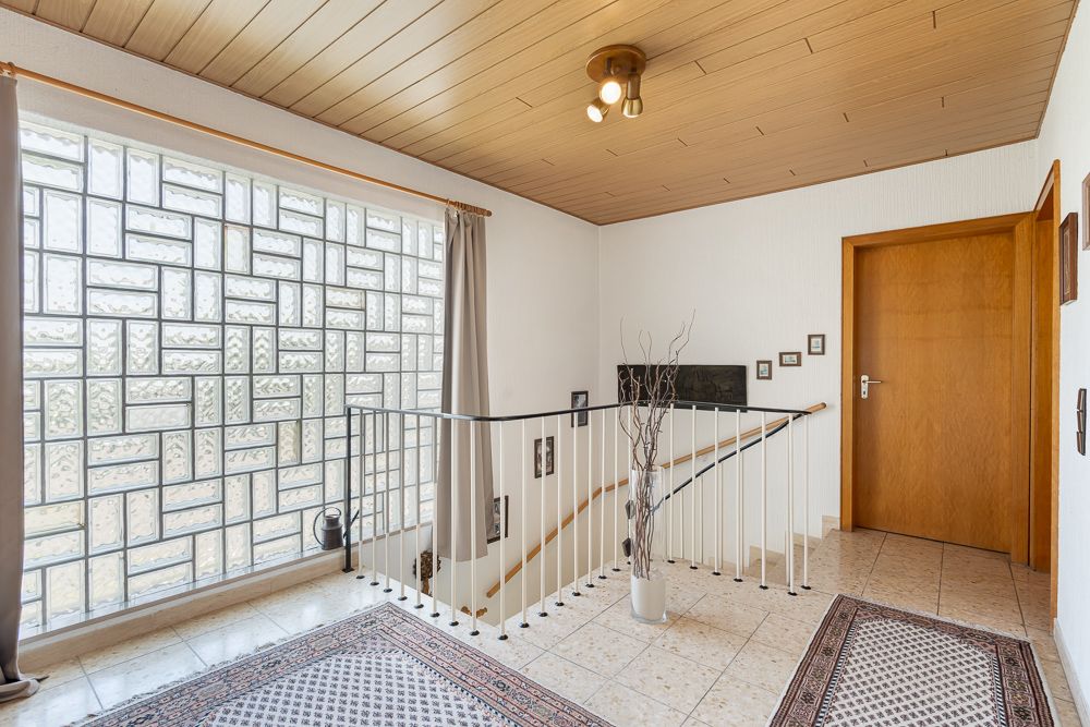 Heller Flur mit Teppichen, Treppengeländer, Glaselement und Holztür.