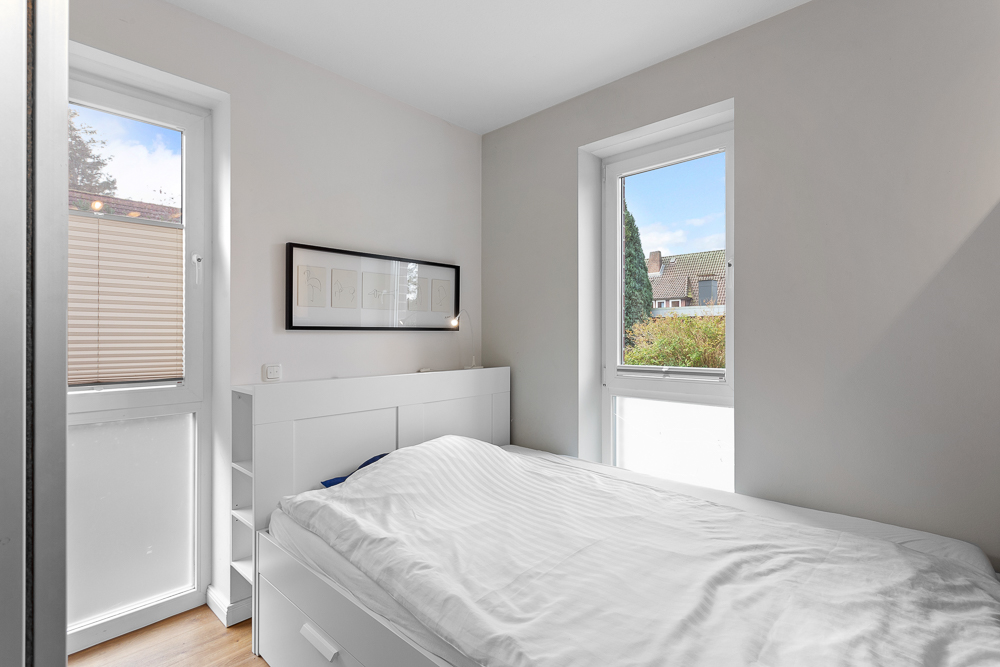 Helles Schlafzimmer mit weißem Bett, zwei Fenstern und einem gerahmten Bild an der Wand.