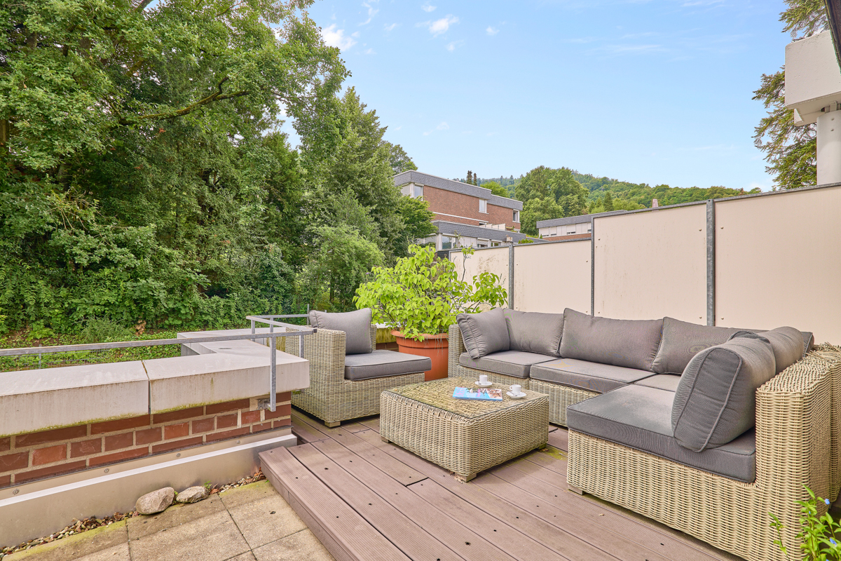 Terrasse mit grauem Rattan-Sofa, Tisch, Pflanze und Blick auf Bäume und Wohnhäuser.
