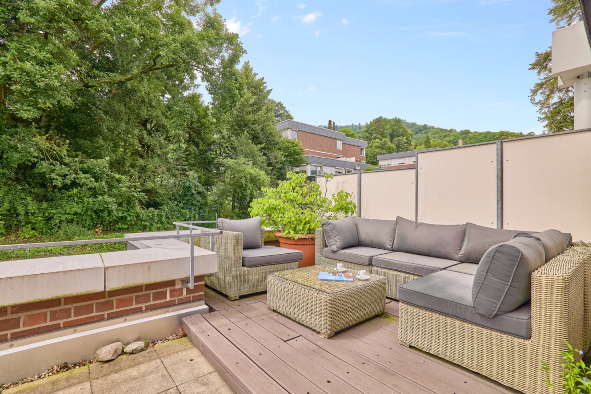 Terrasse mit grauem Rattan-Sofa, Tisch, Pflanze und Blick auf Bäume und Wohnhäuser.
