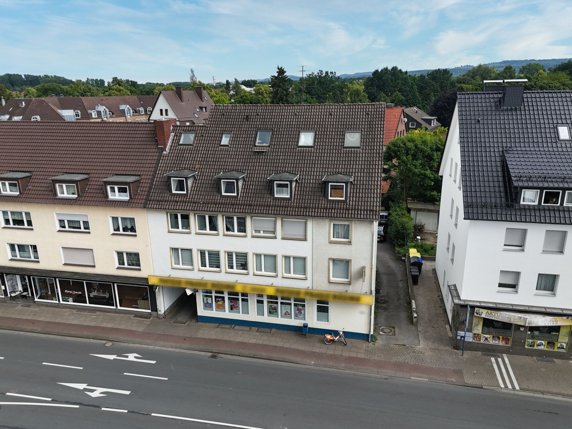 Mehrstöckiges Wohnhaus mit Schaufenstern im Erdgeschoss an einer Straße, daneben Gehweg und Nachbarhäuser.