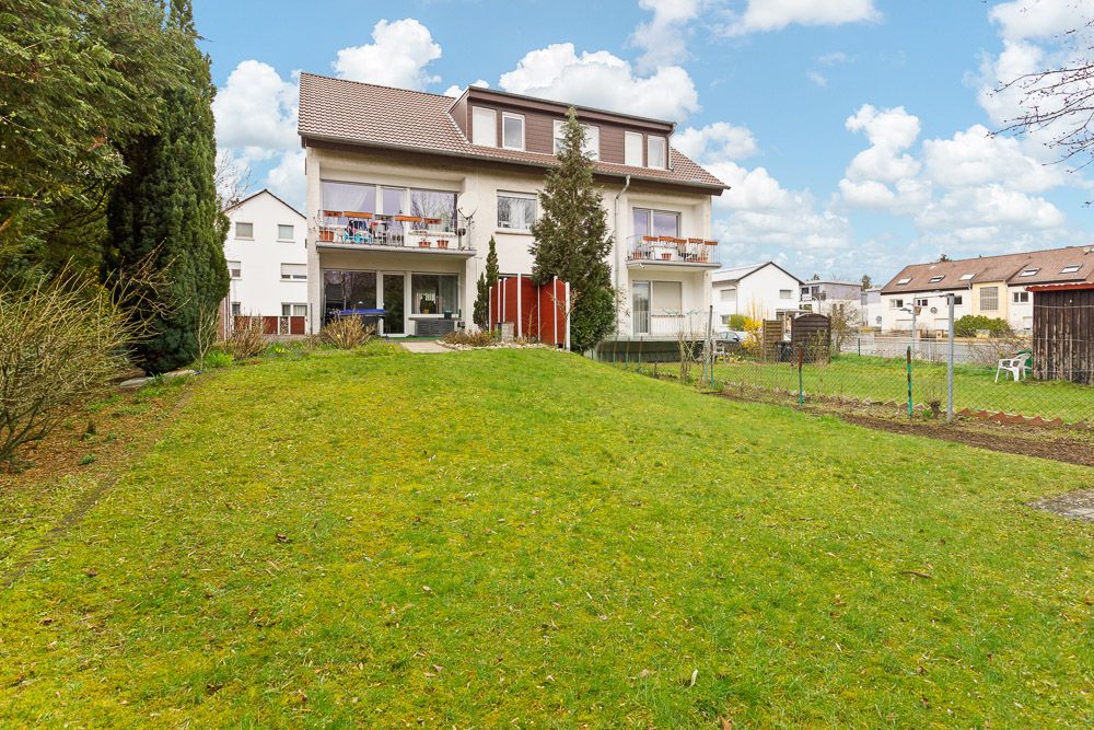 Zweistöckiges Wohnhaus mit Balkon und großem Garten, im Vordergrund eine Rasenfläche.