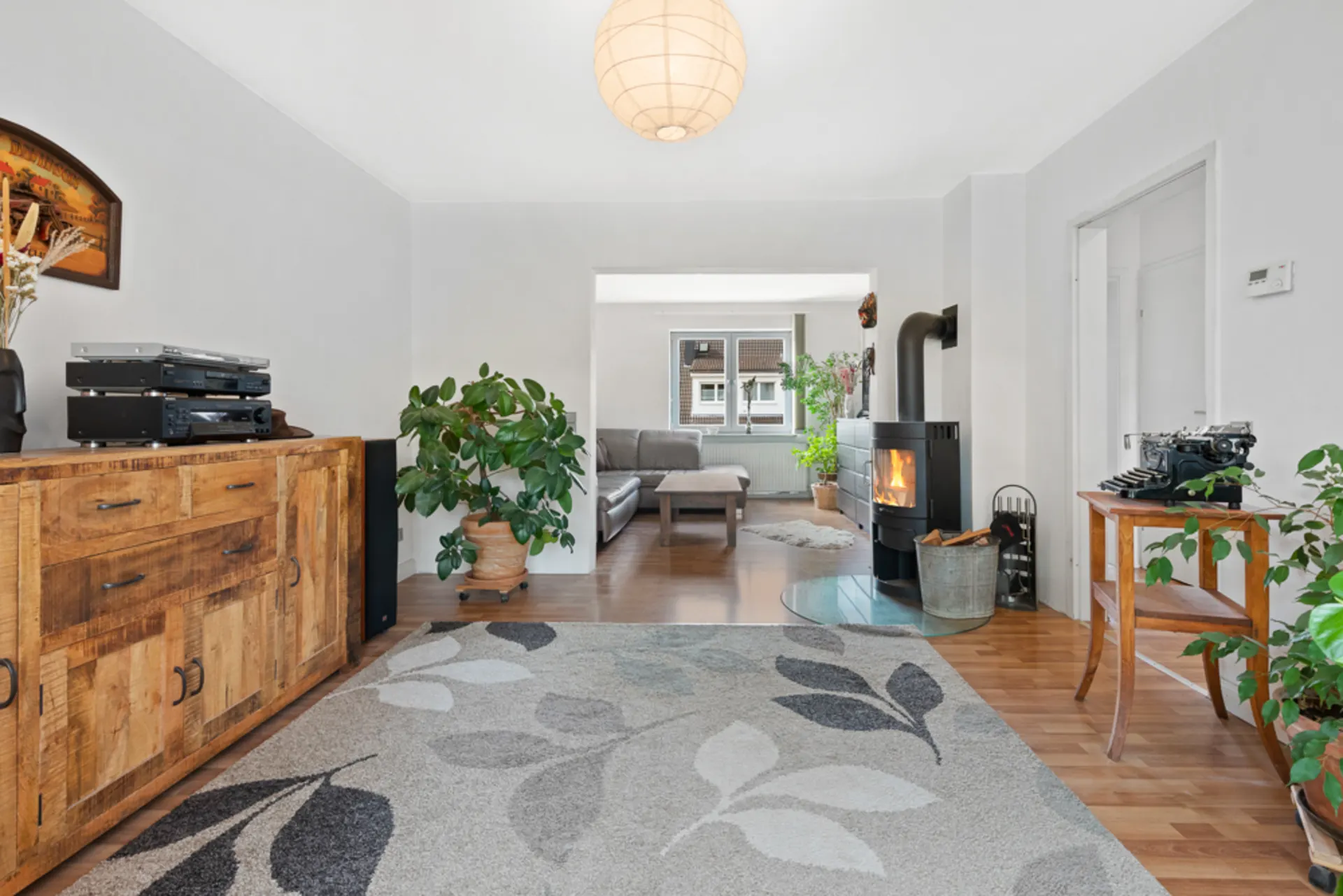 Wohnzimmer mit Holzkommode, Teppich, Kaminofen, Pflanzen und Sofa im Hintergrund.