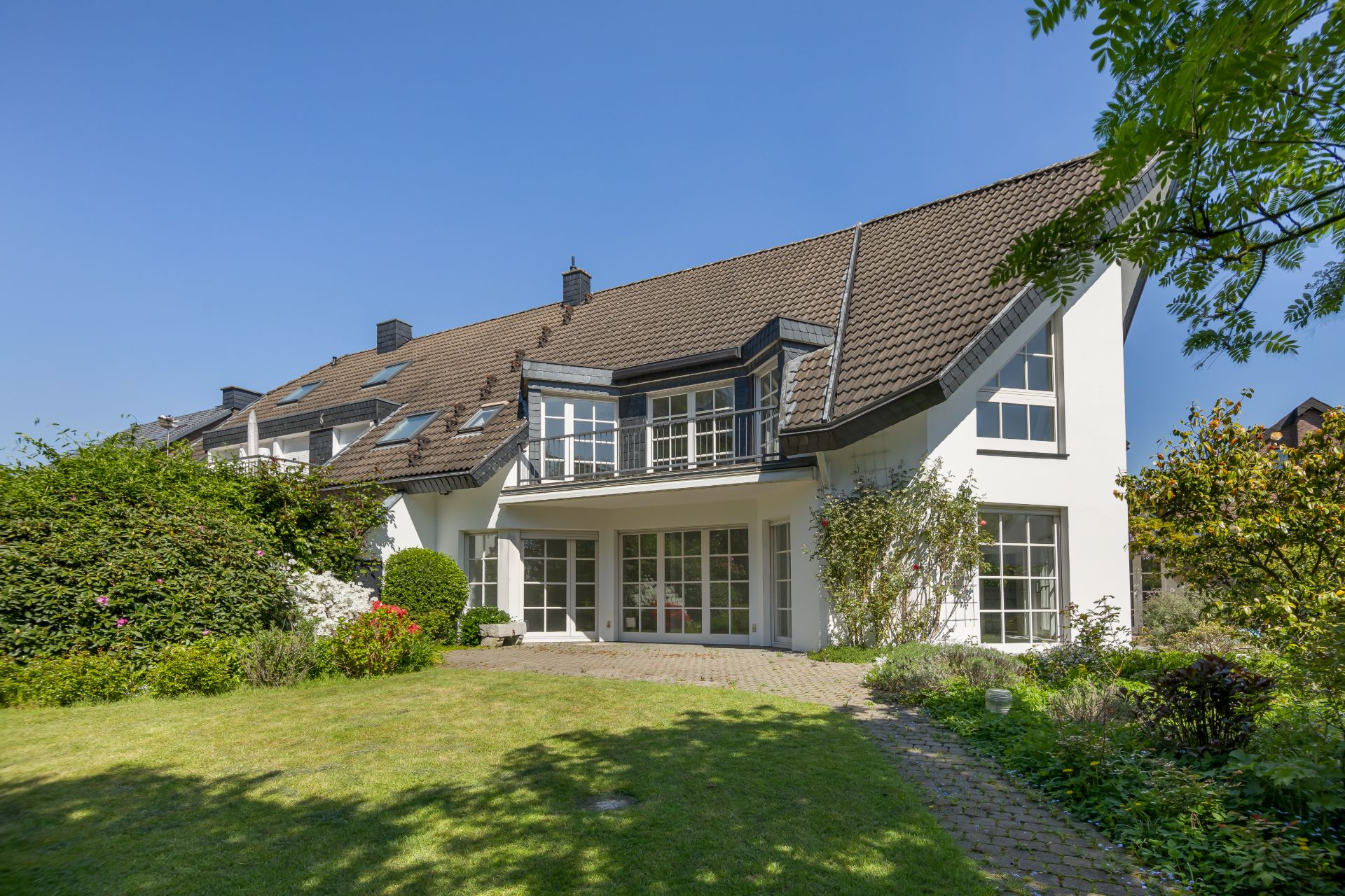 Modernes weißes Wohnhaus mit Garten, großen Fenstern und braunem Dach bei sonnigem Wetter.