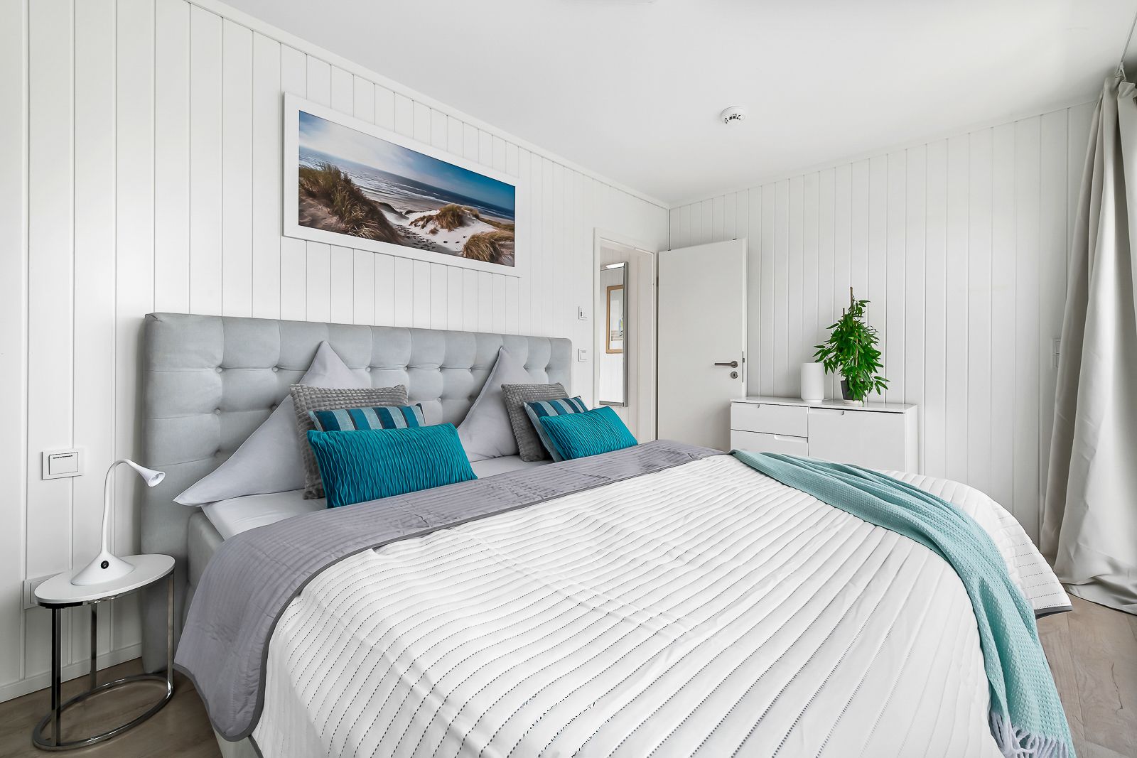 Helles Schlafzimmer mit Doppelbett, weißer Kommode, Pflanze und Bild an der Wand.