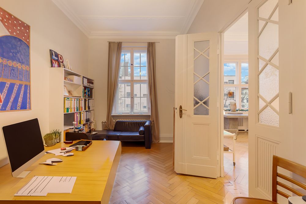 Helles Arbeitszimmer mit Schreibtisch, Regal, Sofa und großem Fenster, offene Tür zu weiterem Raum.