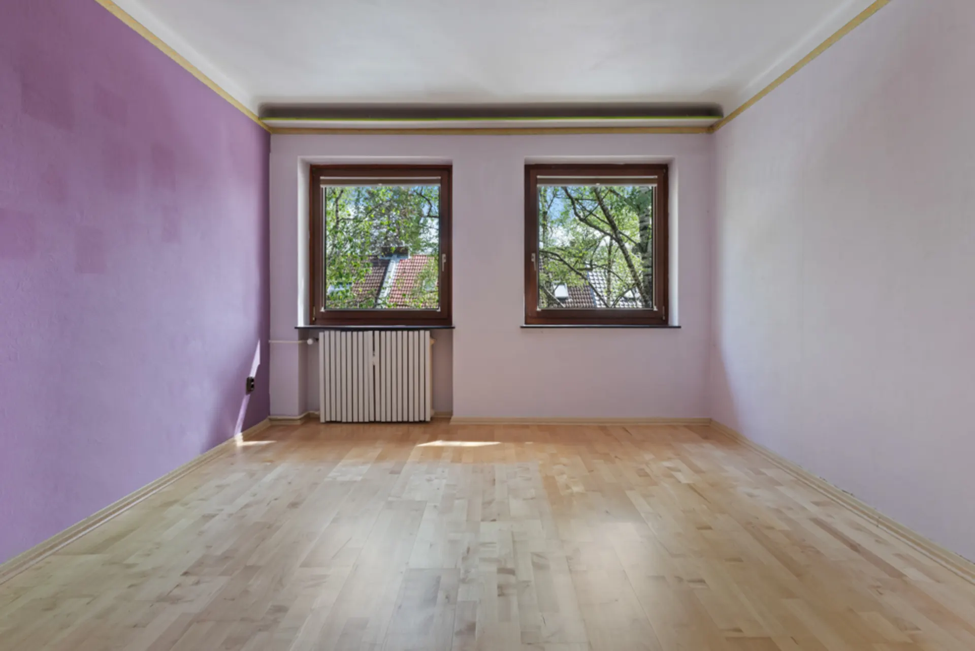 Leeres Zimmer mit hellvioletten Wänden, zwei Fenstern und hellem Holzboden.