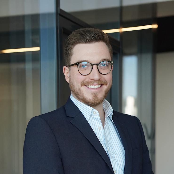 Ein lächelnder Mann mit Brille und Anzug steht vor einer Glaswand.