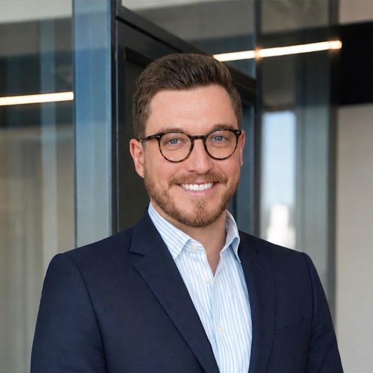 Ein lächelnder Mann mit Brille und Anzug steht vor einer Glaswand.
