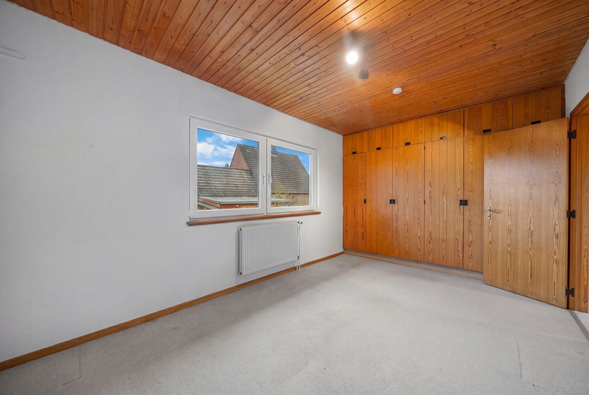 Leeres Zimmer mit Holzdecke, Einbauschrank und großem Fenster mit Blick nach draußen.