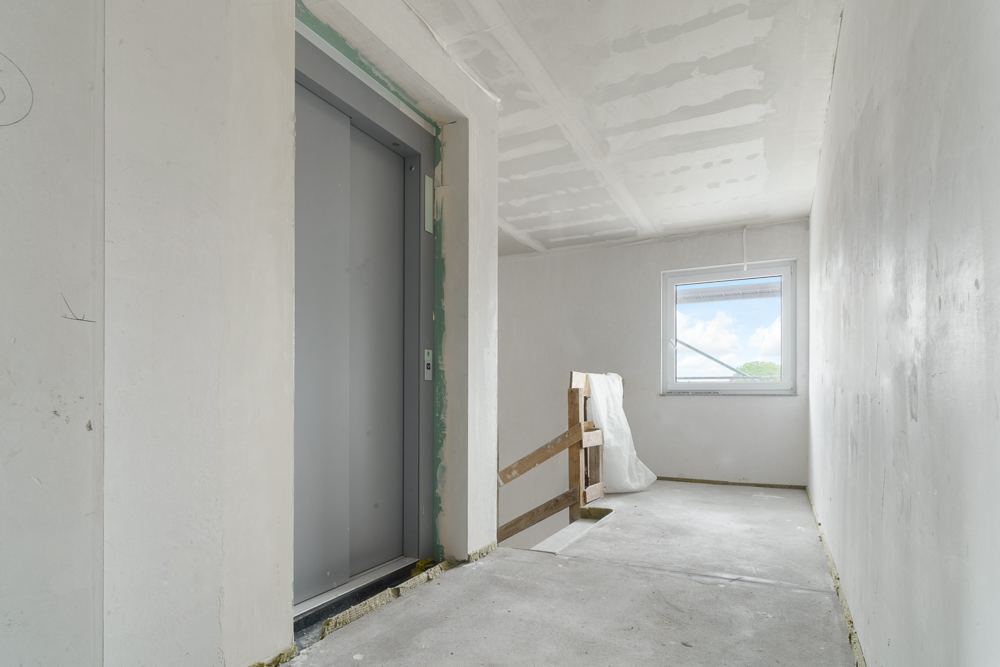 Heller Flur mit Aufzug, Fenster und unfertigem Geländer, Wände im Rohbauzustand.