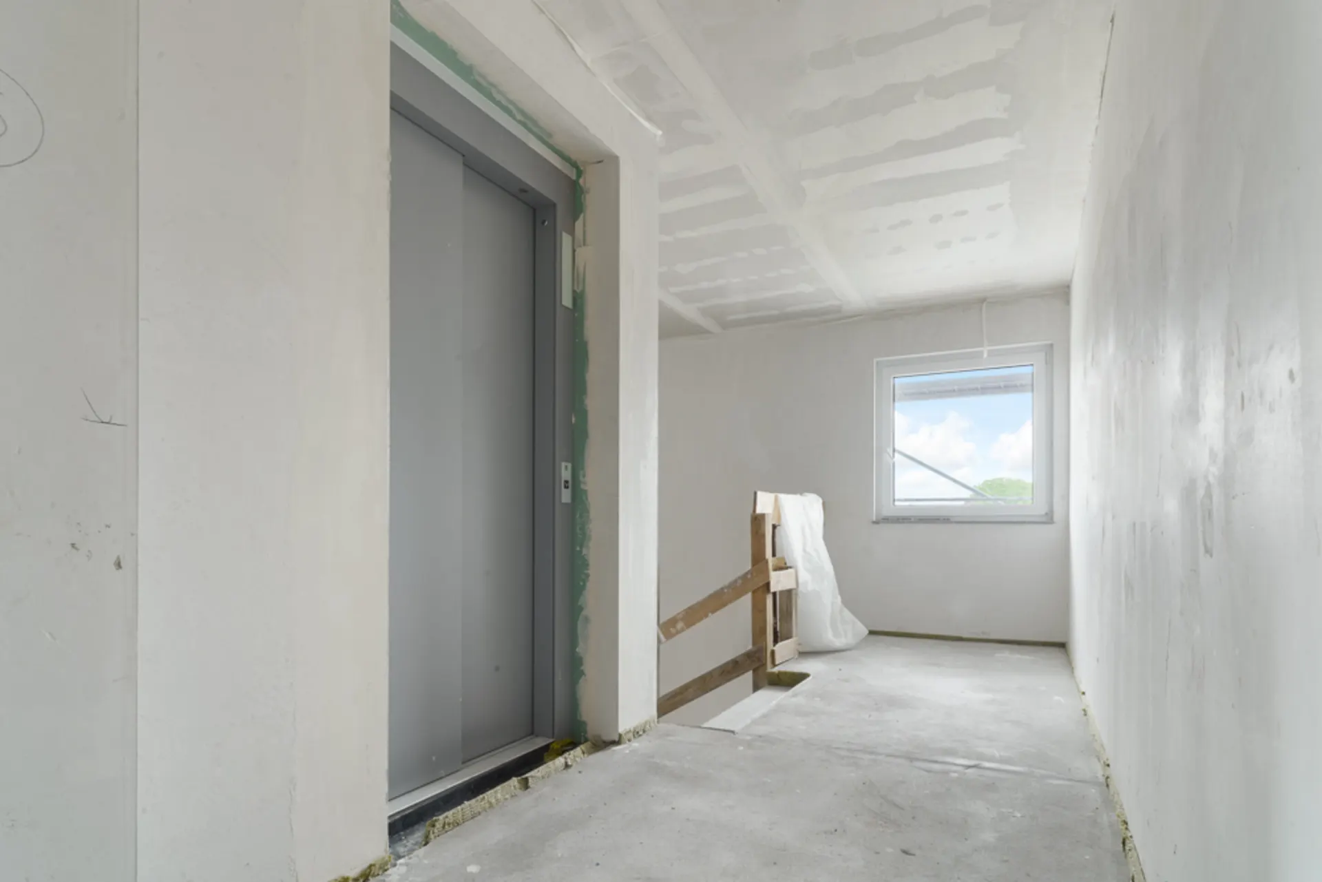Heller Flur mit Aufzug, Fenster und unfertigem Geländer, Wände im Rohbauzustand.