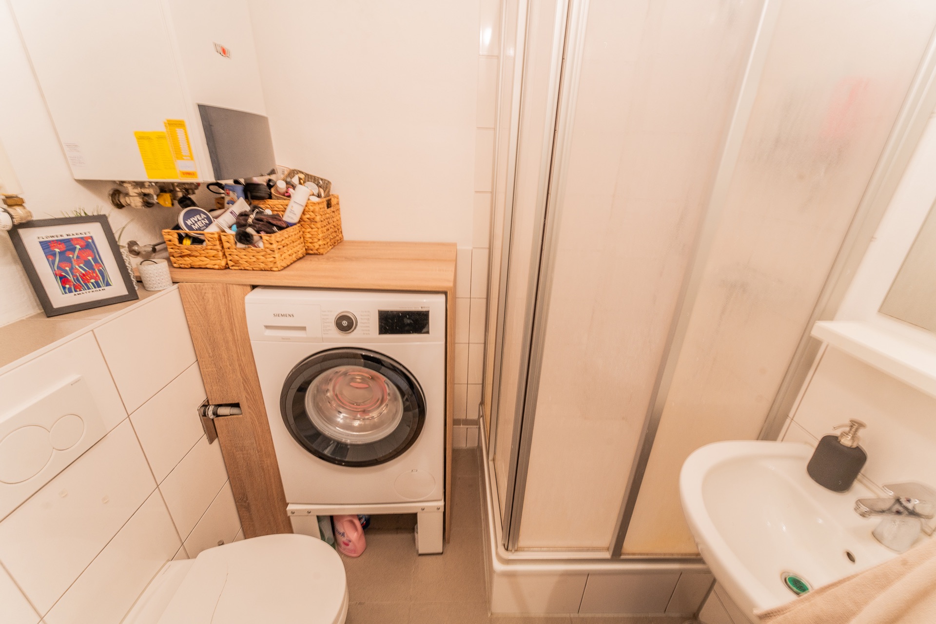 Kleines Badezimmer mit Dusche, Waschmaschine, Toilette, Waschbecken und Kosmetikartikeln.