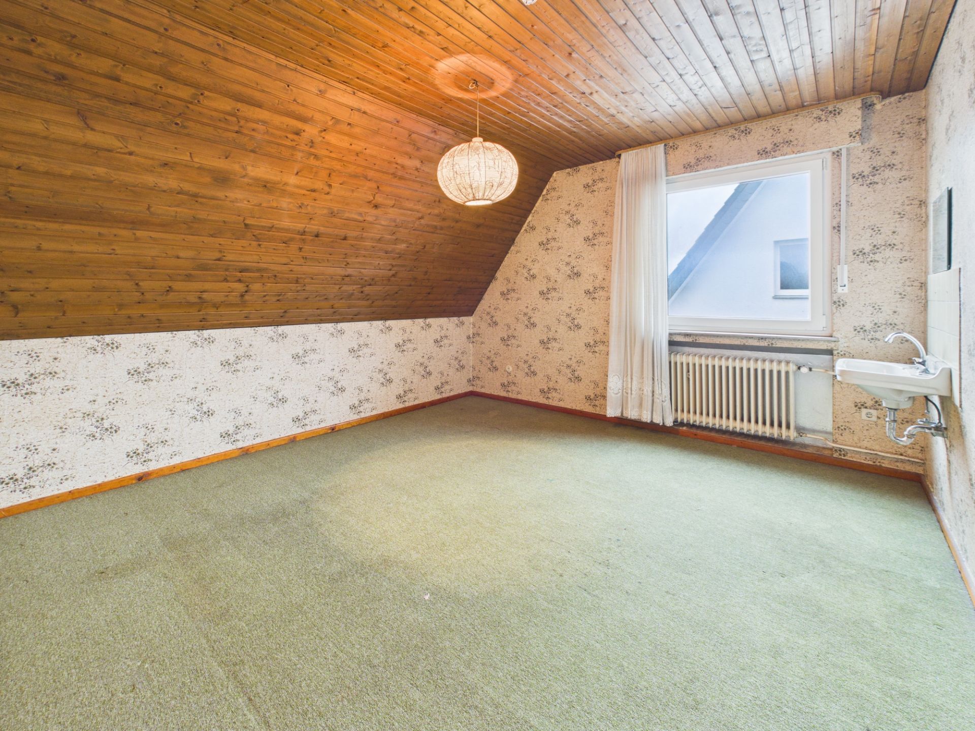 Zimmer mit Teppichboden, Holzdecke, Waschbecken und großem Fenster mit Vorhang.
