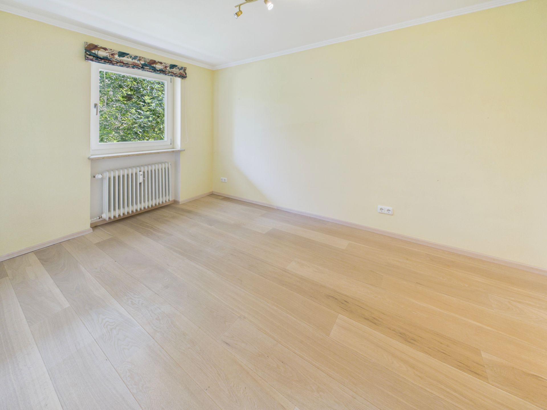 Helles, leeres Zimmer mit Holzfußboden, gelben Wänden und einem Fenster mit Heizkörper.