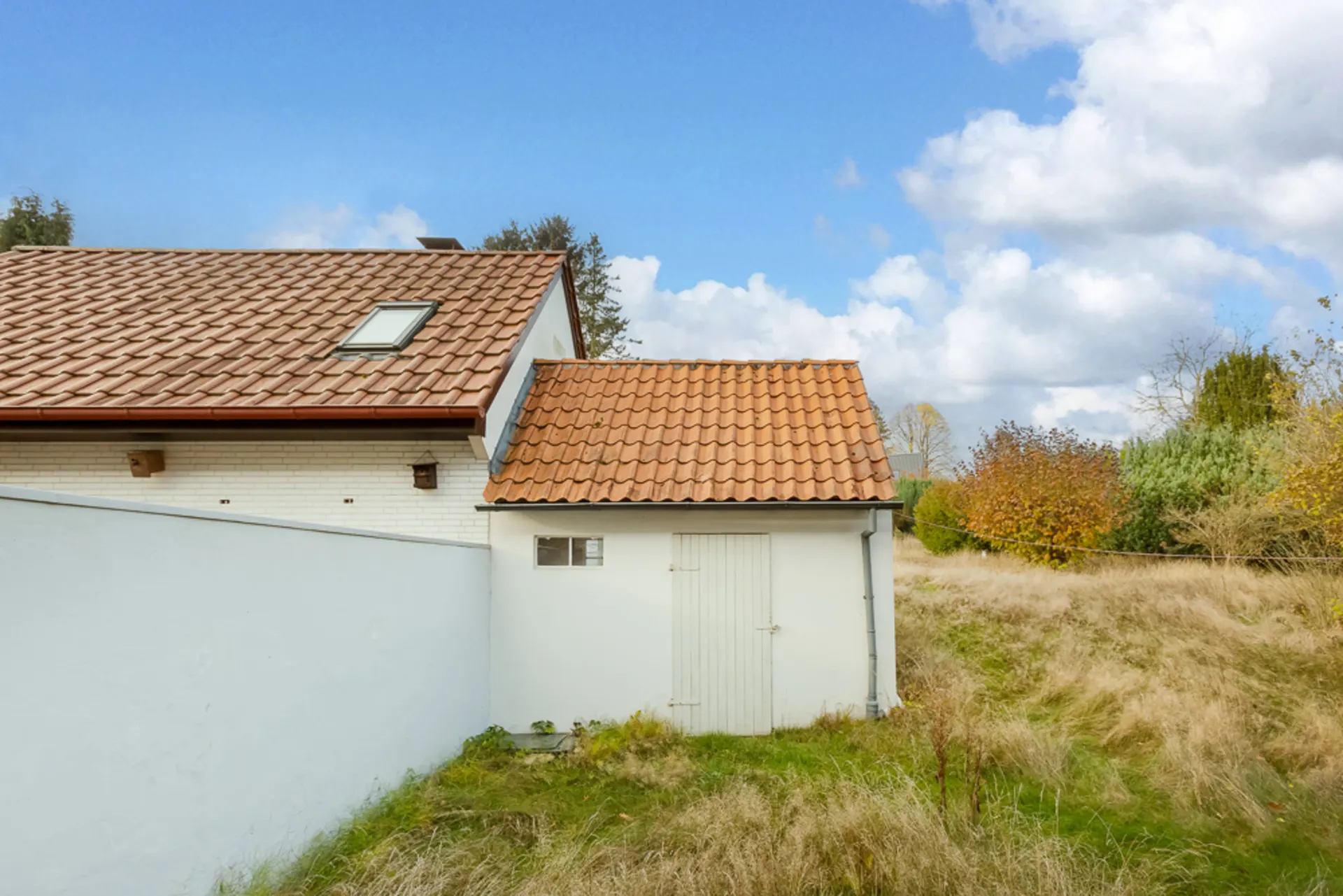 Weißes Nebengebäude mit rotem Ziegeldach neben einem Haus, umgeben von Gras und Bäumen.