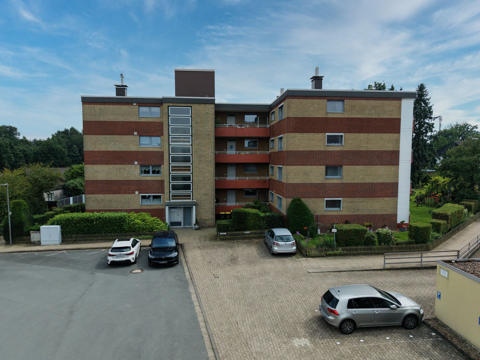 Vierstöckiges Wohnhaus mit roten Streifen, mehrere geparkte Autos davor, grüne Hecken rundherum.
