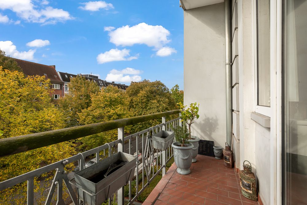 Balkon mit roten Fliesen, Pflanzkübeln und Blick auf Bäume und Gebäude im Hintergrund.