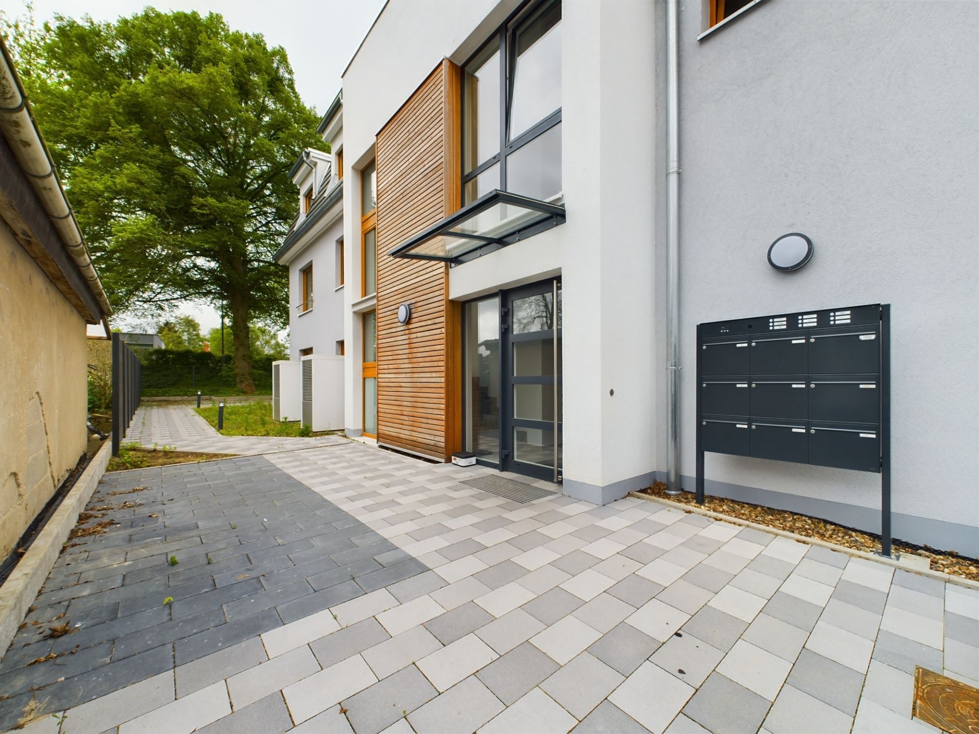 Eingang eines modernen Wohnhauses mit Briefkästen, gepflastertem Weg und großer Glasfront.