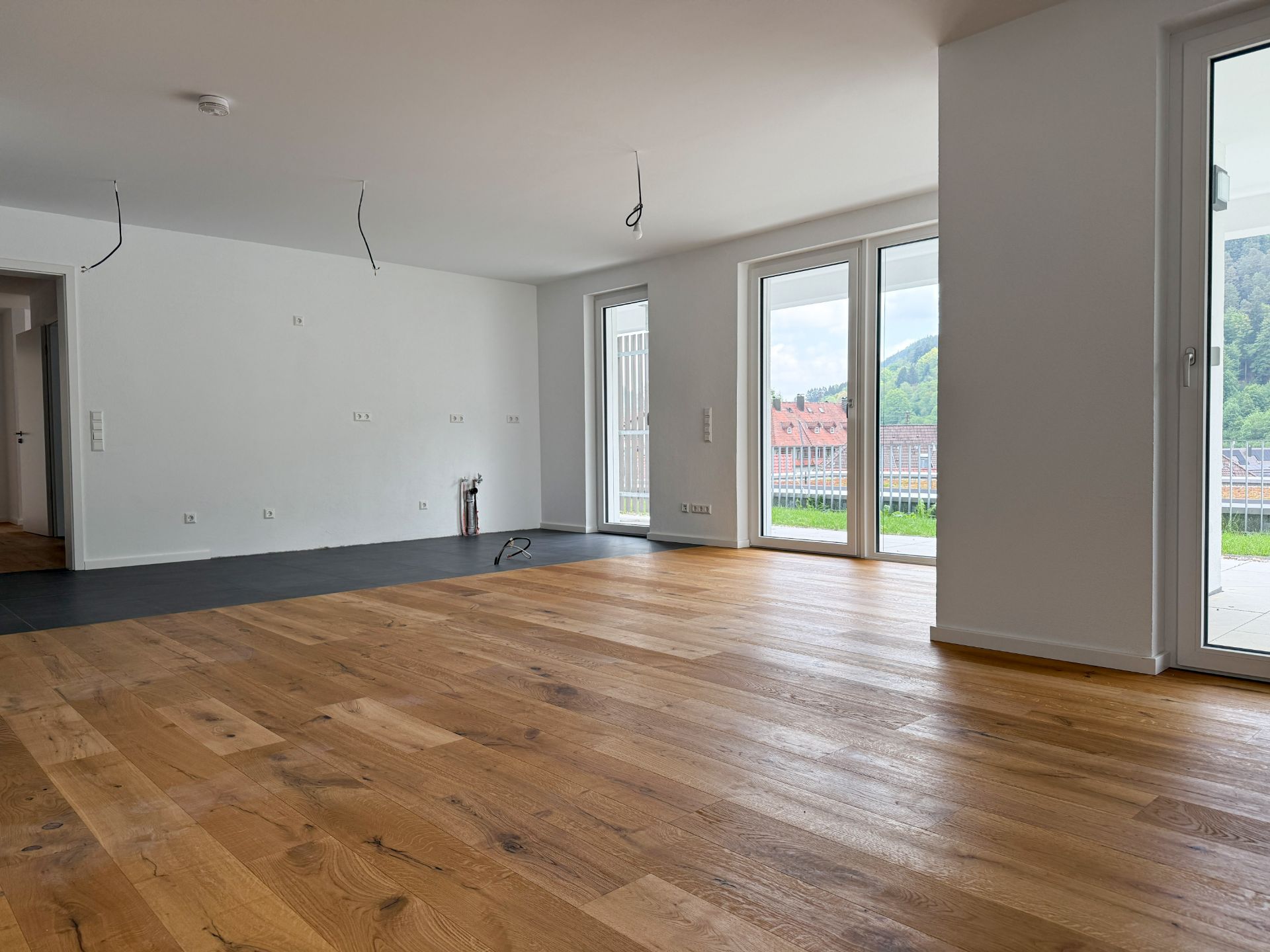 Helles, leeres Zimmer mit Holzfußboden und großen Fenstern, Blick ins Grüne.
