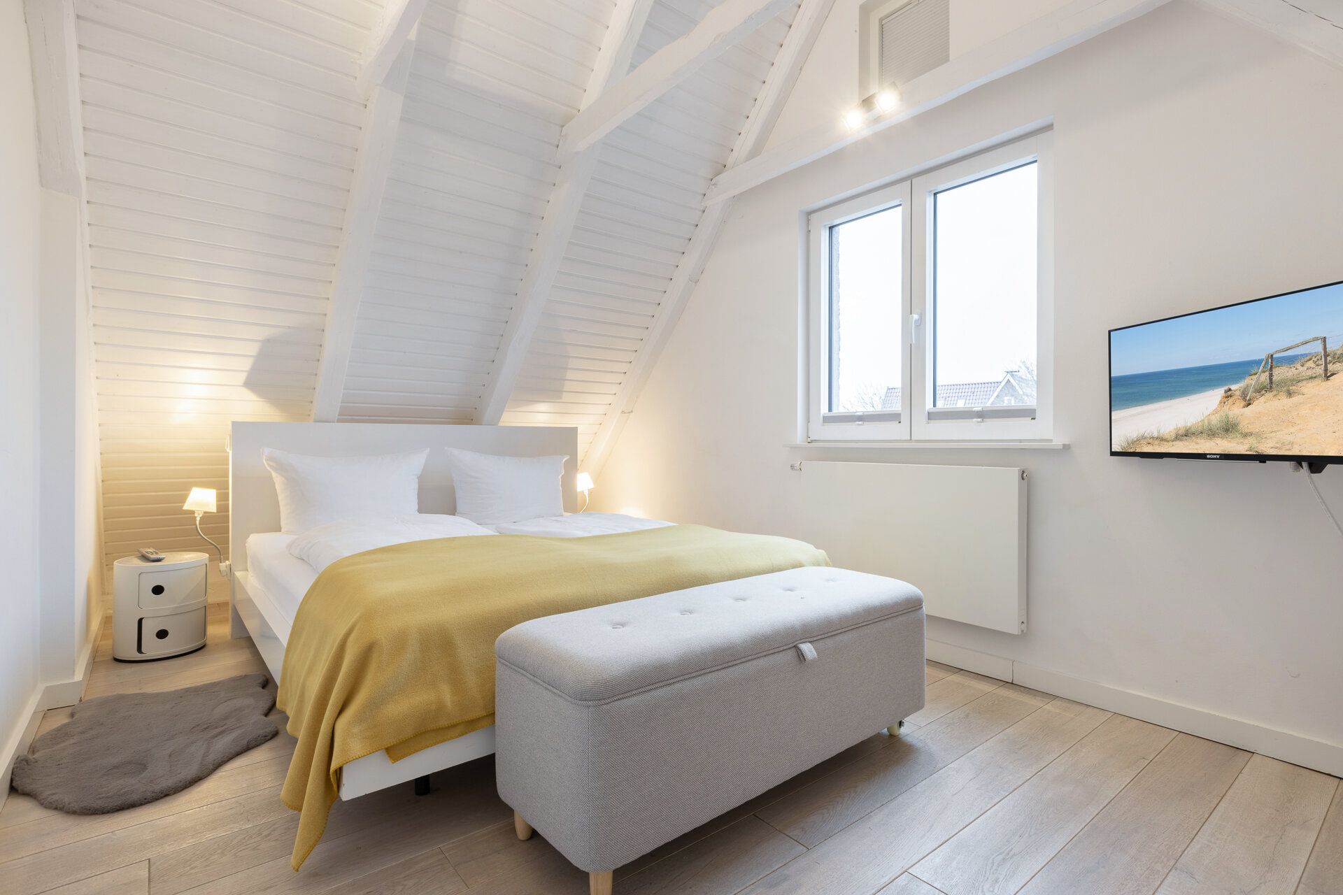 Helles Schlafzimmer mit Doppelbett, gelber Decke, Fenster und Fernseher an der Wand.