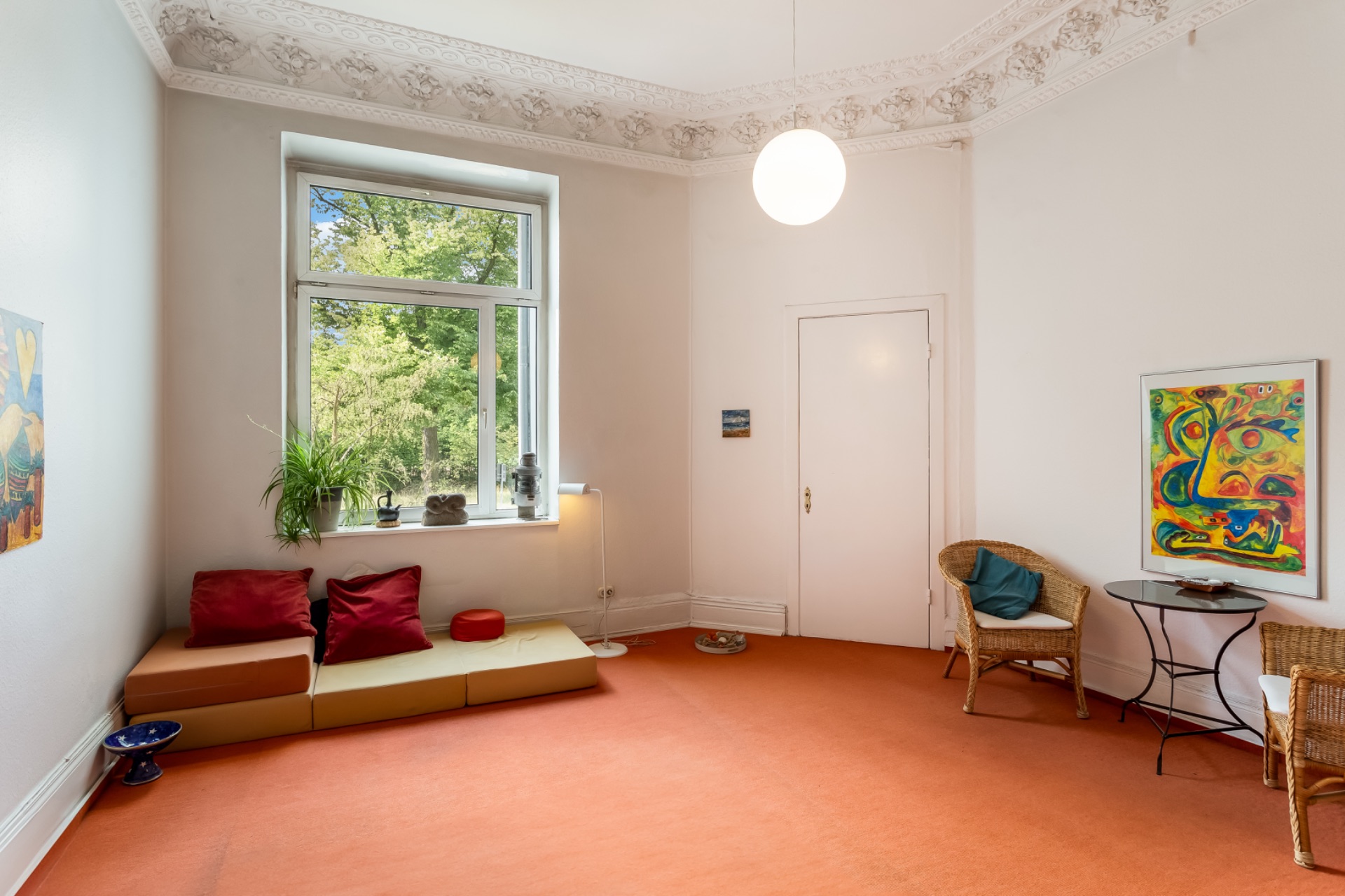 Helles Wohnzimmer mit orangefarbenem Teppich, Fenster, Sesseln, kleinem Tisch und Wanddekoration.