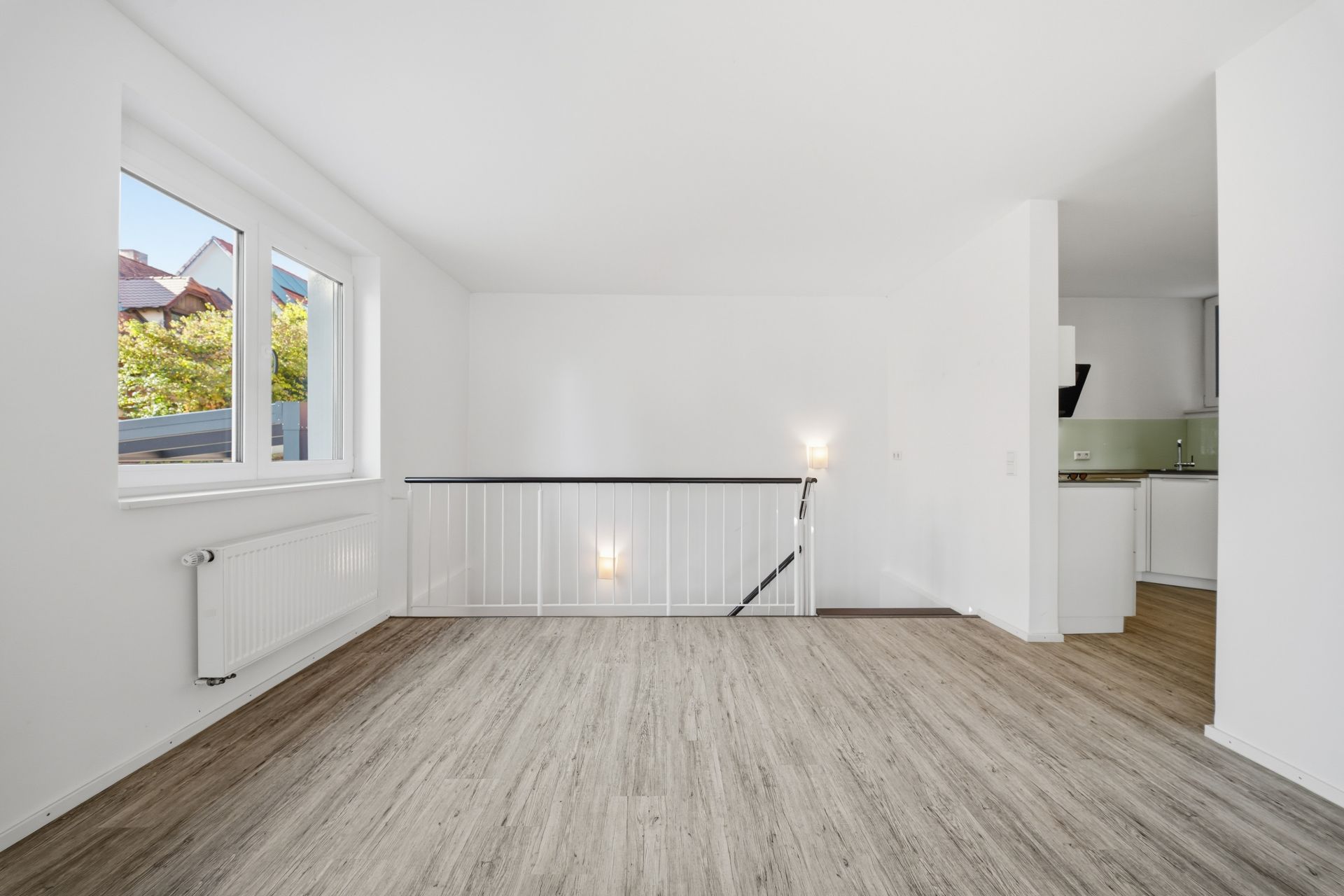 Heller Raum mit Holzboden, Fenster, Treppengeländer und Blick in eine offene Küche.