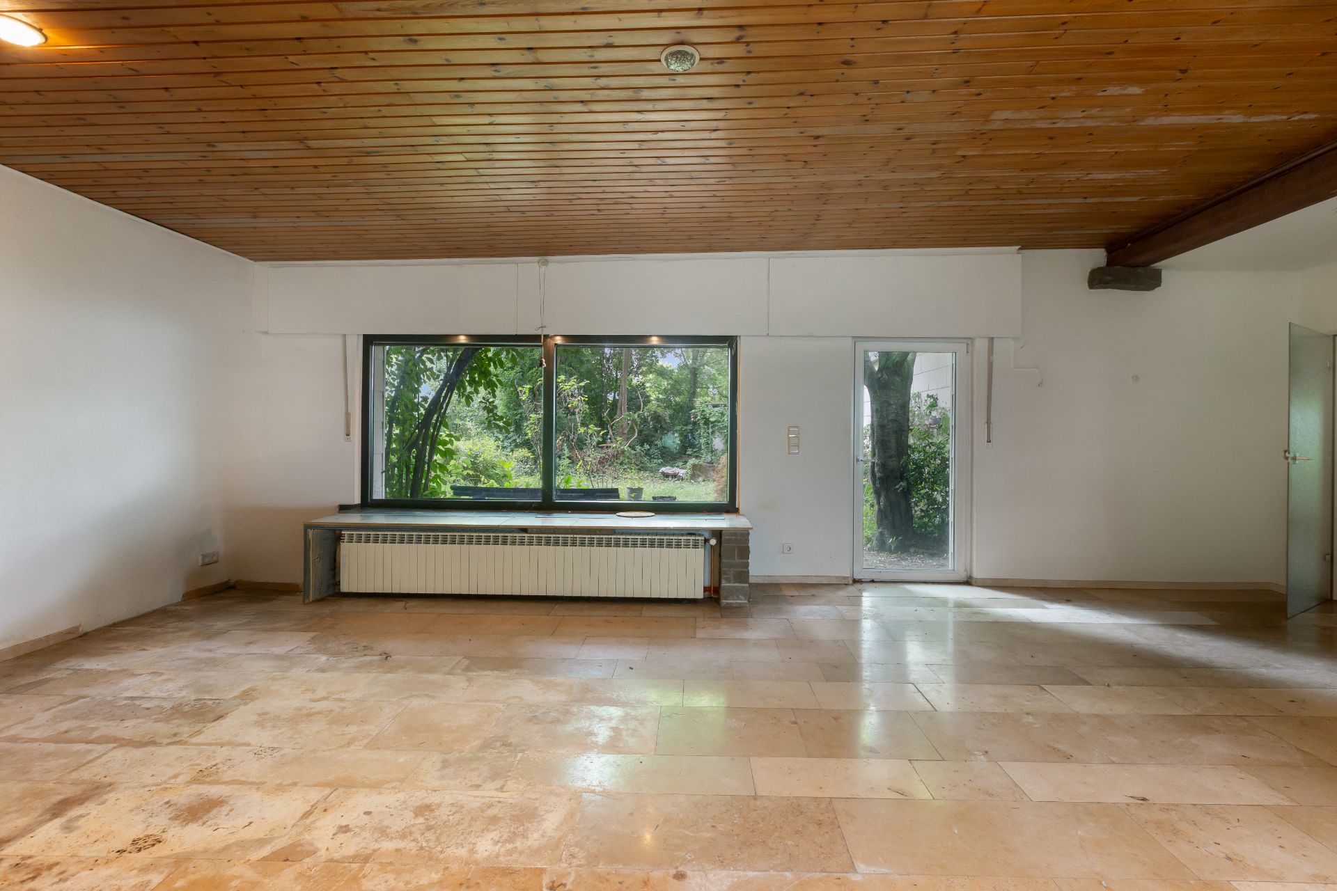 Leeres Zimmer mit großem Fenster, Holzbalkendecke und hellem Fliesenboden, Blick in den Garten.