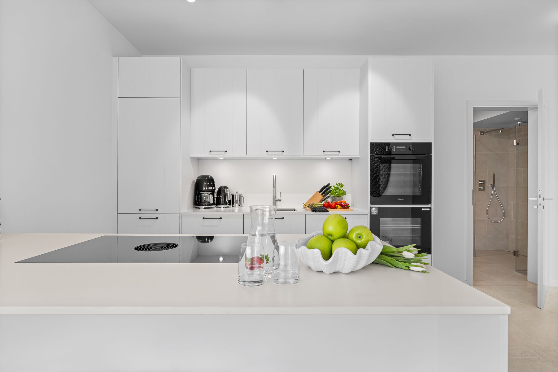 Moderne weiße Küche mit Kochinsel, Äpfeln, Glaskaraffen und Blick ins Badezimmer.