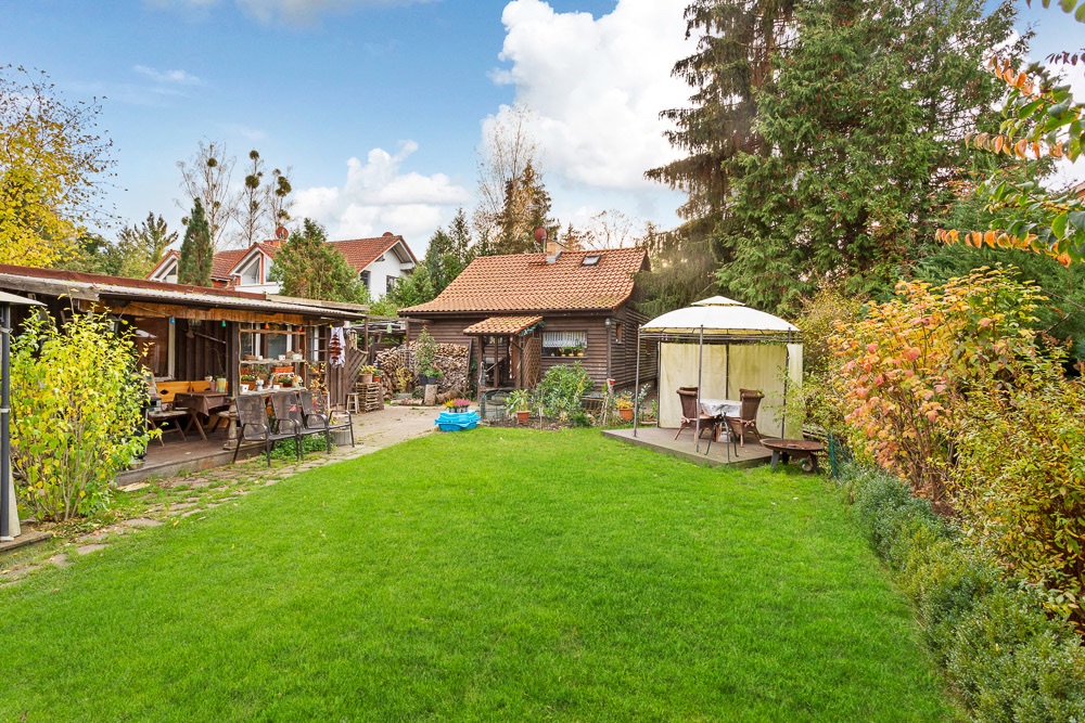 Großer Garten mit Rasen, Holzhaus, überdachter Terrasse und Pavillon mit Sitzmöbeln.