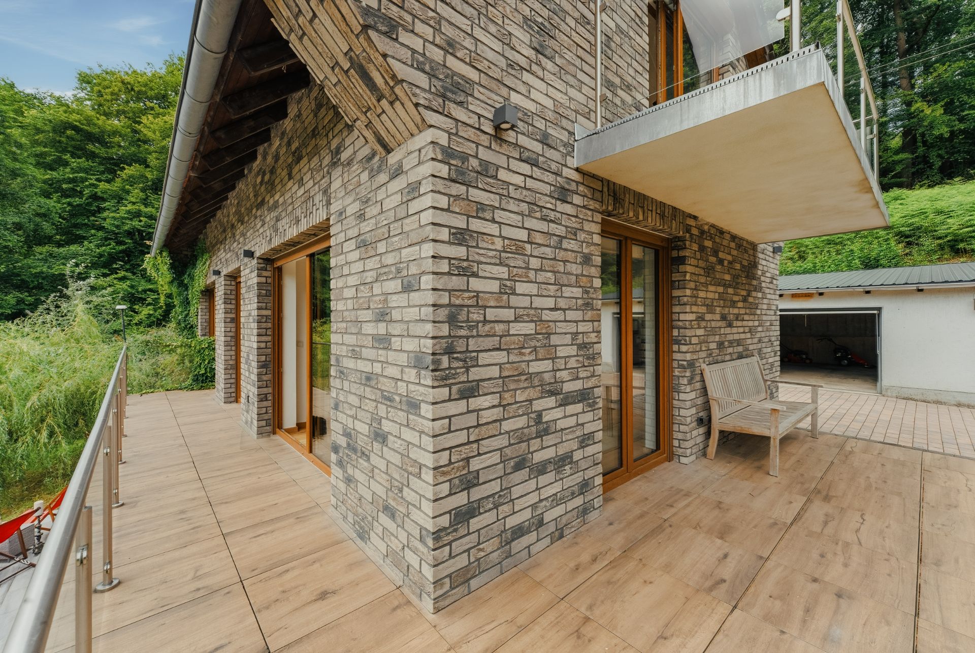 Eckansicht eines Hauses mit Klinkerfassade, Holzbalkon und Gartenbank auf der Terrasse.