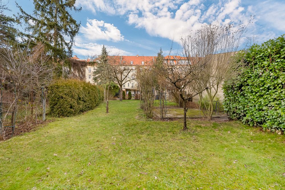 Grüner Garten mit Bäumen und Sträuchern, im Hintergrund ein Wohnhaus mit rotem Dach.