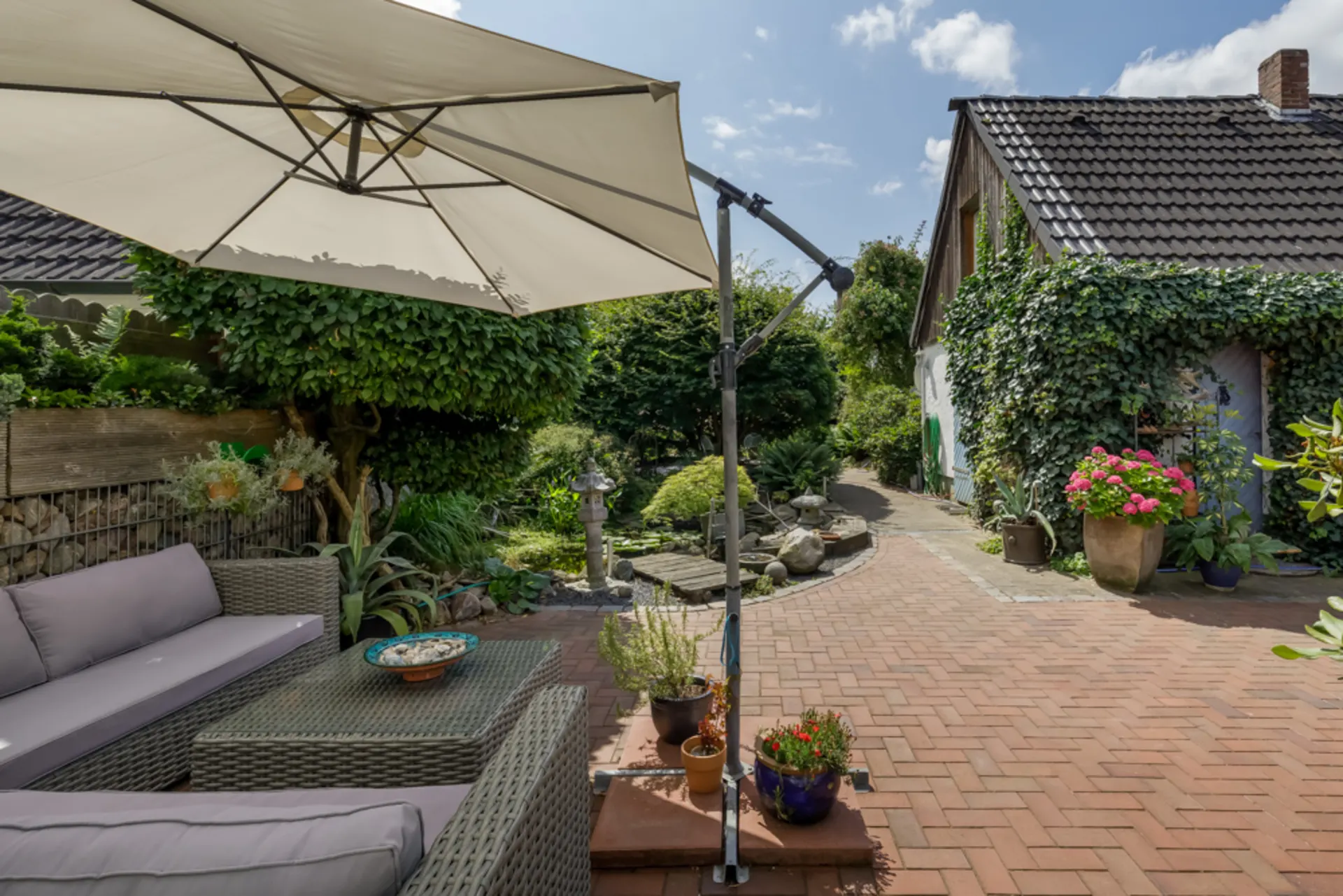 Gepflasterte Terrasse mit Sitzgruppe, Sonnenschirm und bepflanztem Garten neben einem Haus.