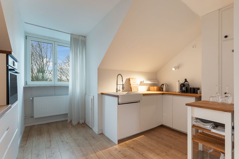 Helle Einbauküche mit Holzboden, weißen Schränken und großem Fenster mit Gardine.