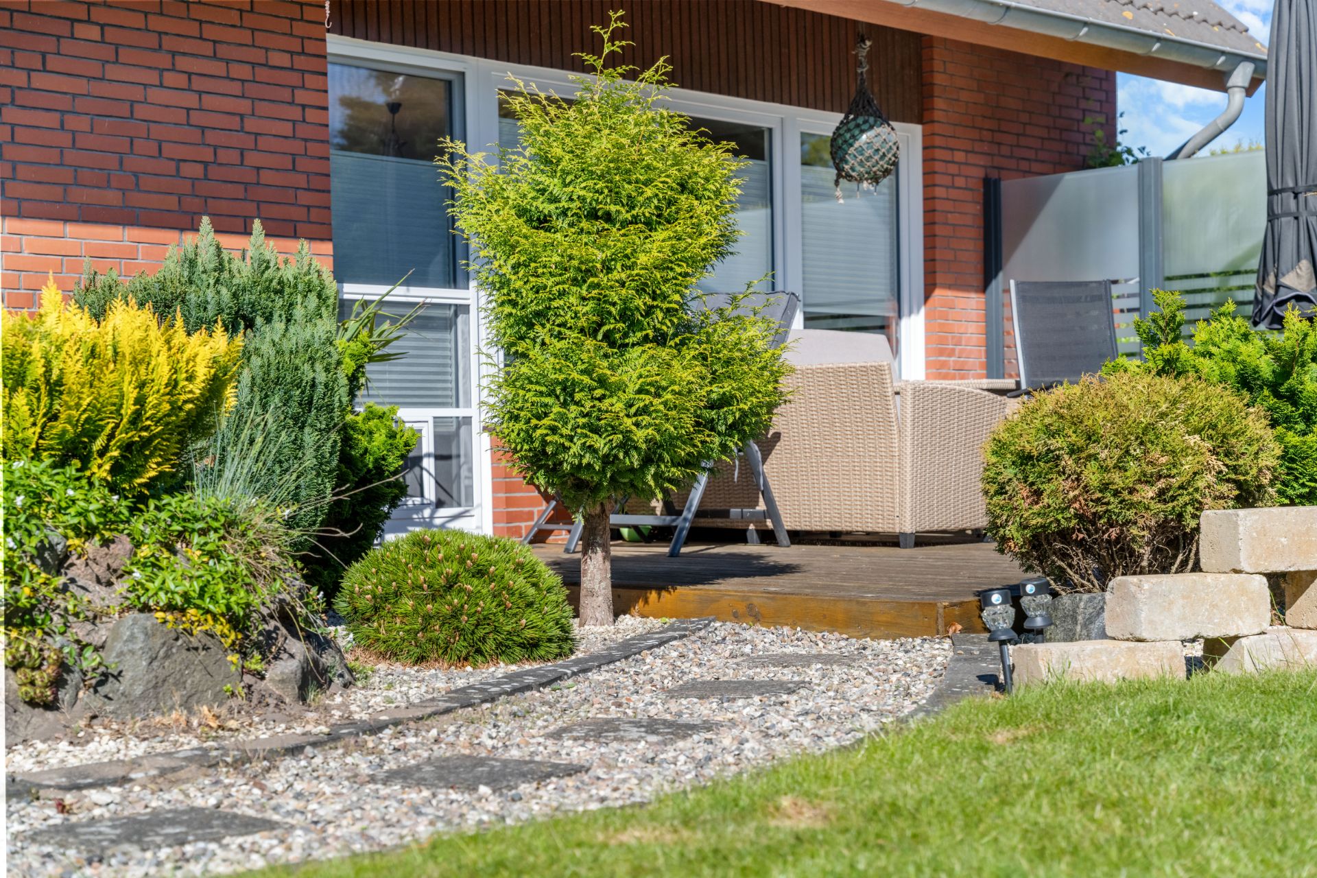 Gepflegter Garten mit Terrasse, Gartenmöbeln, Sträuchern und Steinen vor einem Wohnhaus.