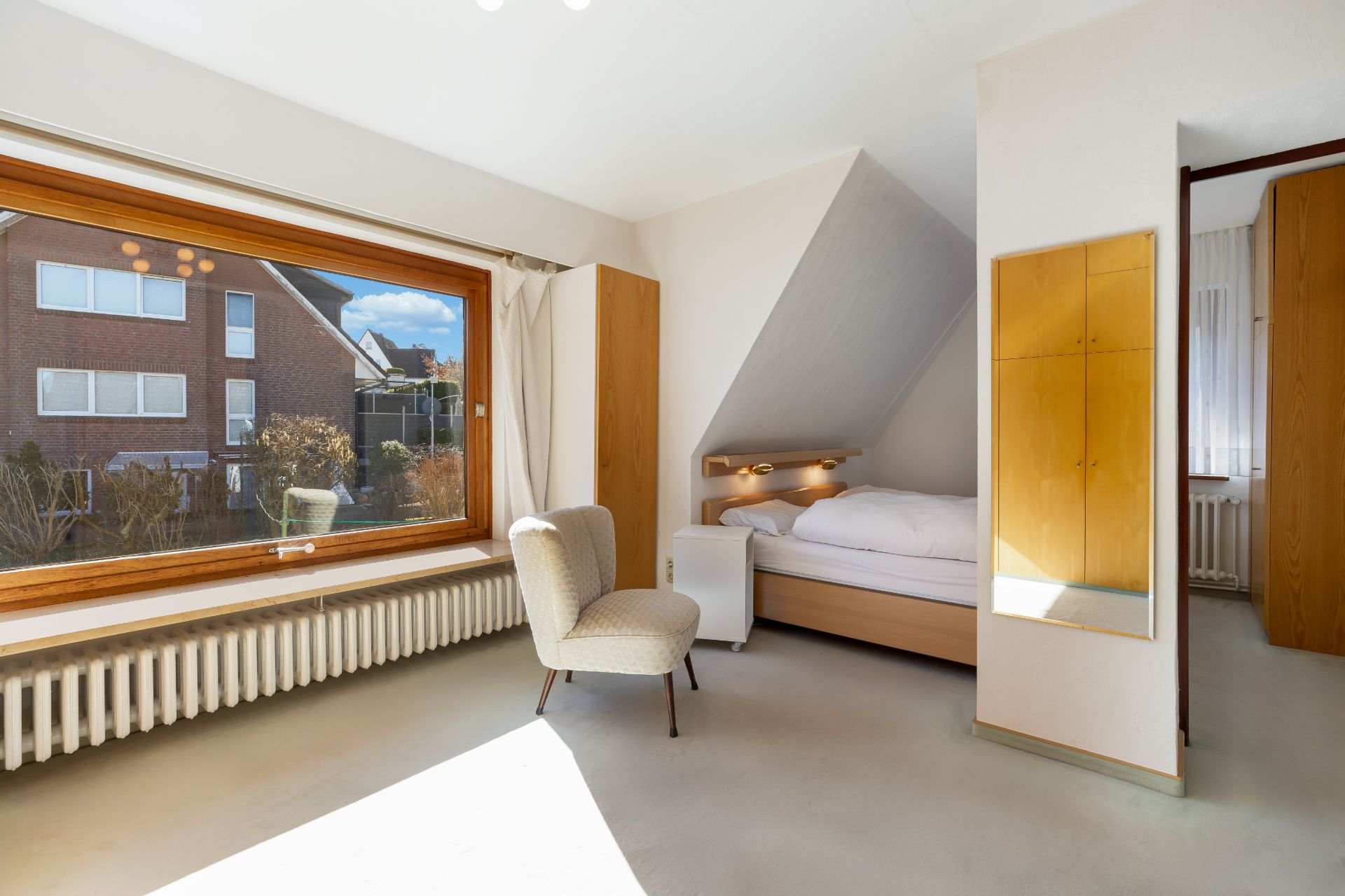 Helles Schlafzimmer mit großem Fenster, Sessel, Bett und Holzmöbeln.